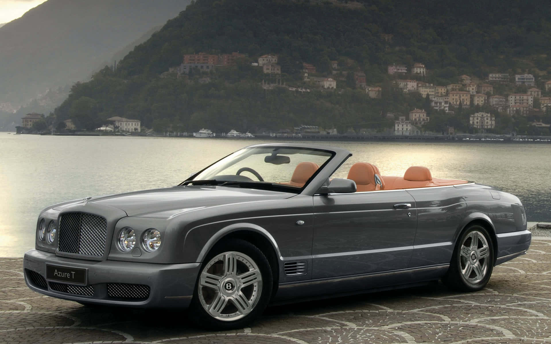 Bentley Azure 桌布 壁紙 — 一架時尚的銀色賓利 Azure T 開篷車，配備棕色真皮座椅，停泊在平靜湖泊旁的石碼頭上，背景是山坡村莊。