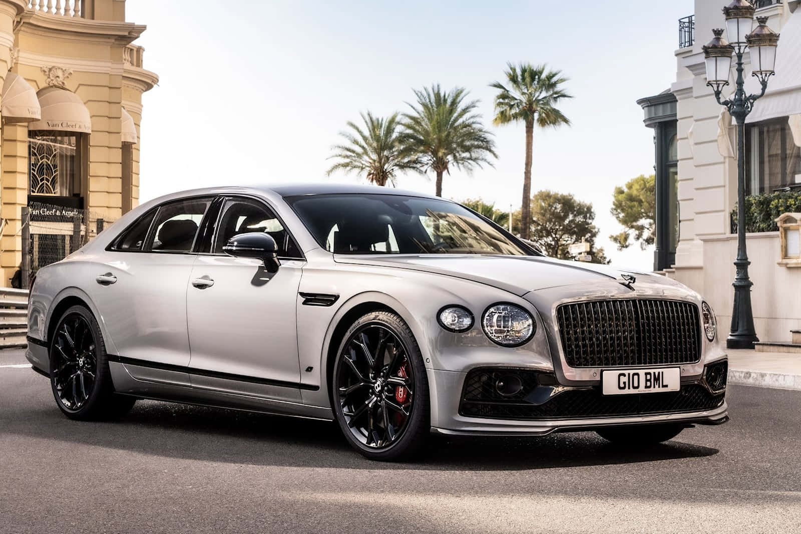 Bentley Flying Spur 桌布 壁紙 — 一架時尚的銀色賓利飛馳，帶有黑色飾邊，停泊在陽光普照的街道上，兩旁種滿棕櫚樹和優雅的建築物。