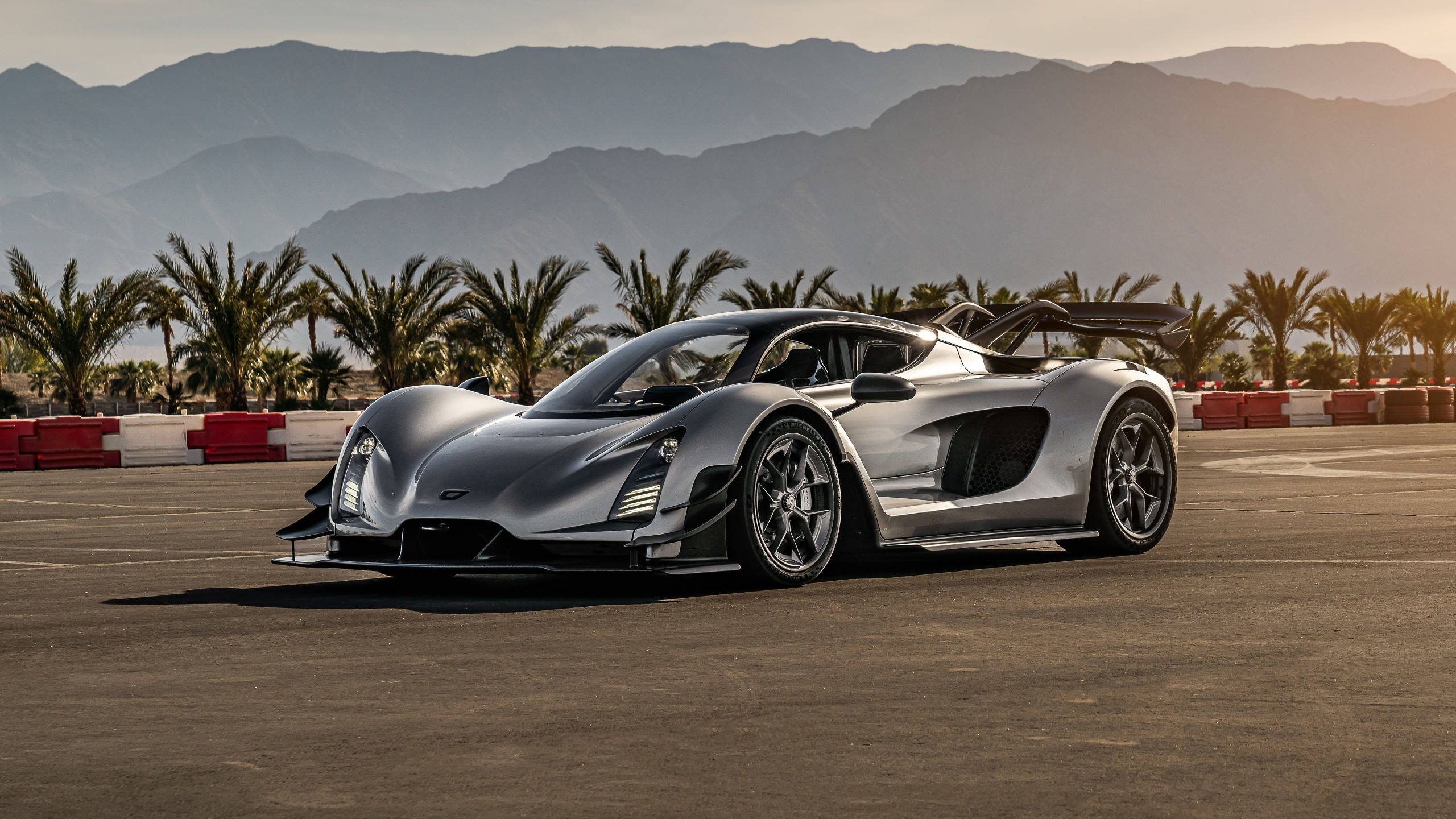 Sports Car 桌布 壁紙 — 一架時尚的銀色超級跑車停在賽道上，背景有棕櫚樹和山脈，夕陽西下。