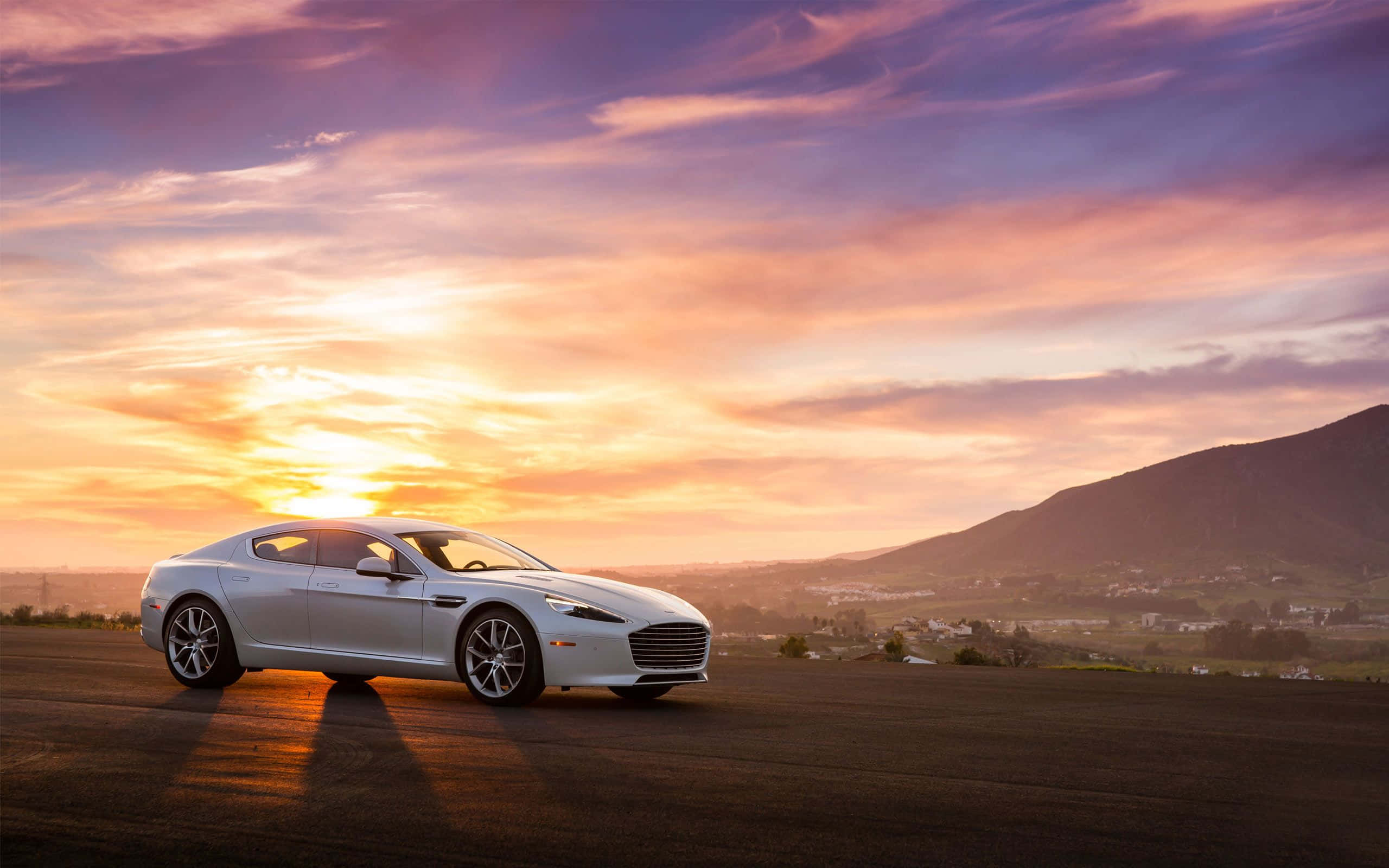 Aston Martin Rapide S 桌布 壁紙 — 一架時尚的銀色跑車停泊在山頂上，夕陽西下，遠山之上有鮮豔的橙色和紫色雲彩。