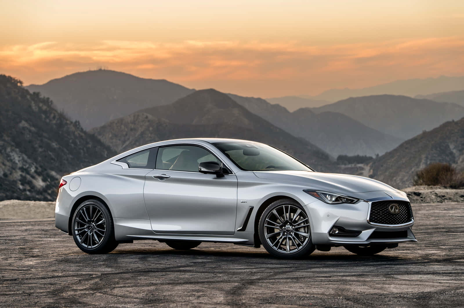Infiniti Q60 桌布 壁紙 — 一架時尚的銀色跑車停泊在日落時分的山頂觀景點，天空呈現橙色，山巒層疊。