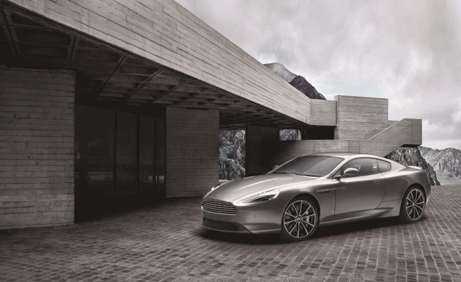 Aston Martin Db9 桌布 壁紙 — 一架時尚的銀色跑車停泊在鵝卵石庭院旁，旁邊是現代粗獷主義混凝土建築，背景有山脈。