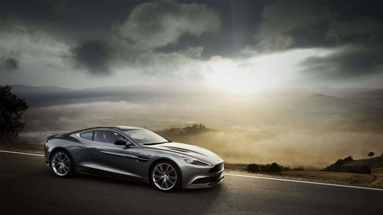 Aston Martin Vanquish 桌布 壁紙 — 一架時尚的銀色跑車在戲劇性的多雲天空下，沿著蜿蜒的山路行駛，陽光穿透雲層。