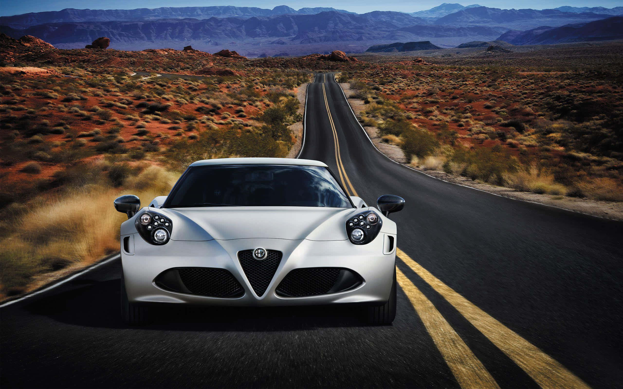 Alfa Romeo 4c 桌布 壁紙 — 一架時尚的銀色跑車在晴朗藍天下，行駛在兩旁紅色岩層的筆直沙漠公路上，車頭朝前。