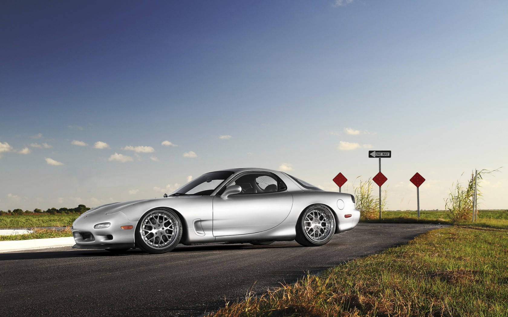 Mazda 桌布 壁紙 — 一架時尚的銀色跑車在晴朗藍天和零星白雲下，沿鄉村道路行駛。