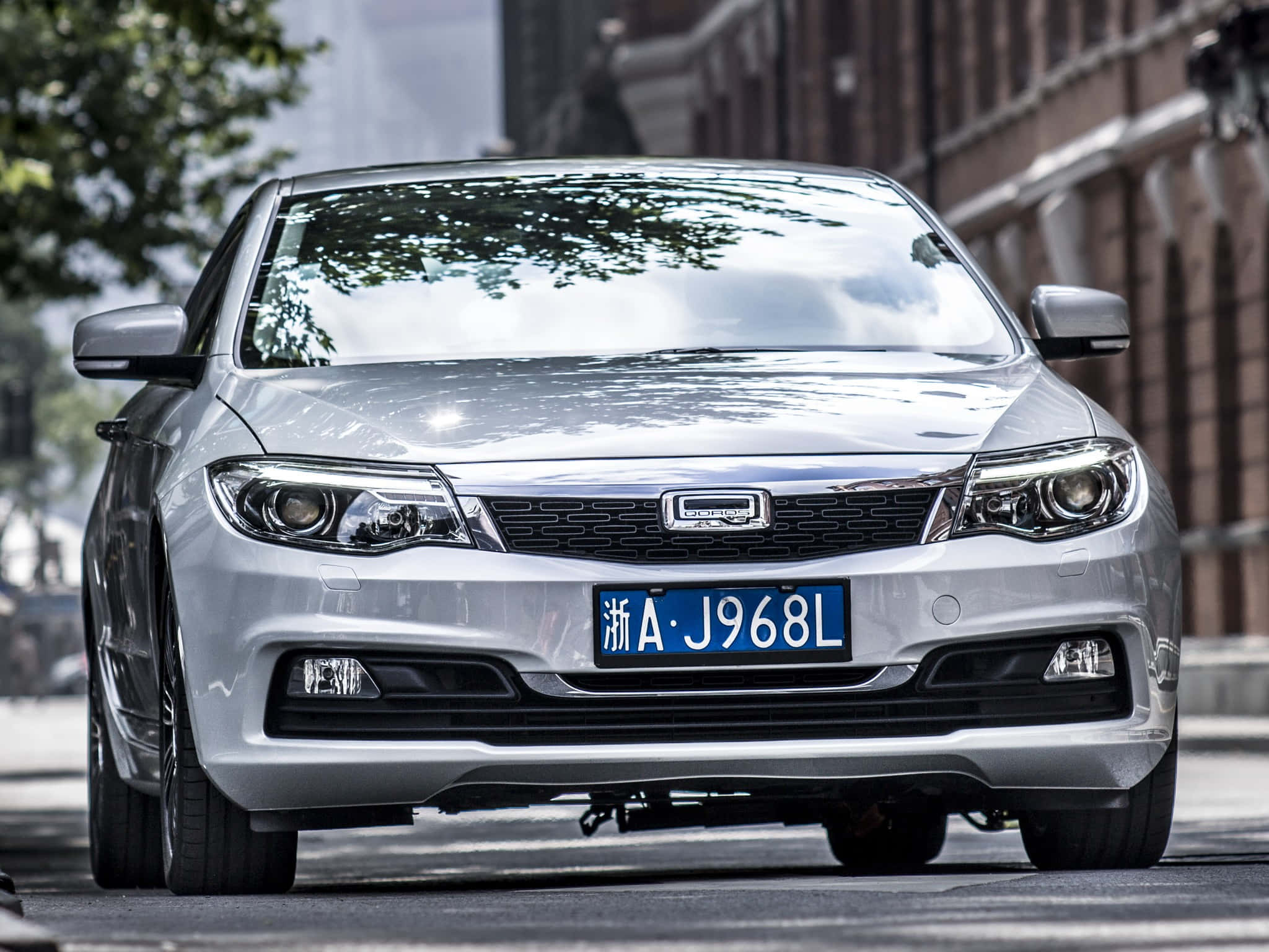 Qoros 桌布 壁紙 — 一架時尚的銀色轎車停泊在城市的街道上，背景有樹木和磚砌建築。