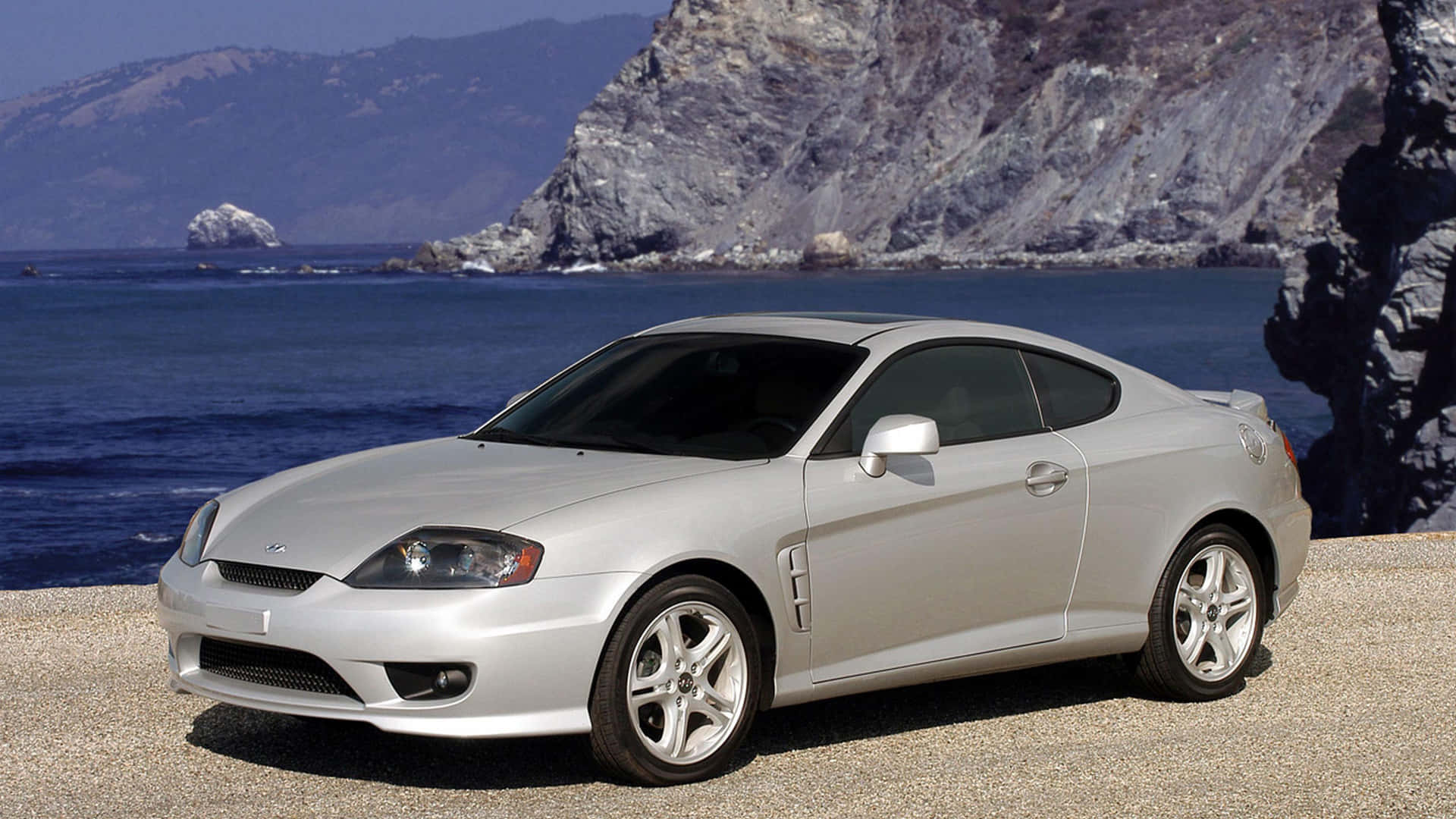Hyundai Tiburon 桌布 壁紙 — 一架時尚的銀色運動轎跑車停在岩石海岸懸崖上，俯瞰深藍色的海洋和崎嶇的懸崖。