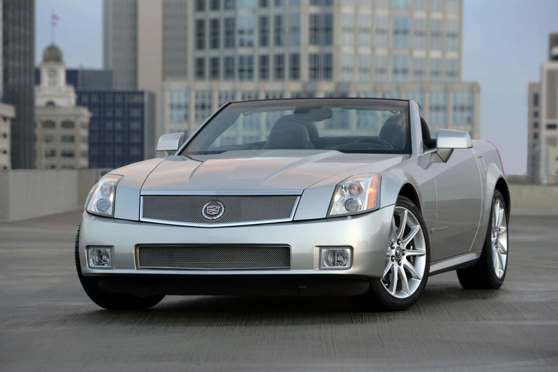 Cadillac Xlr 桌布 壁紙 — 一架時尚的銀色開篷跑車停泊在屋頂上，背景是城市天際線。