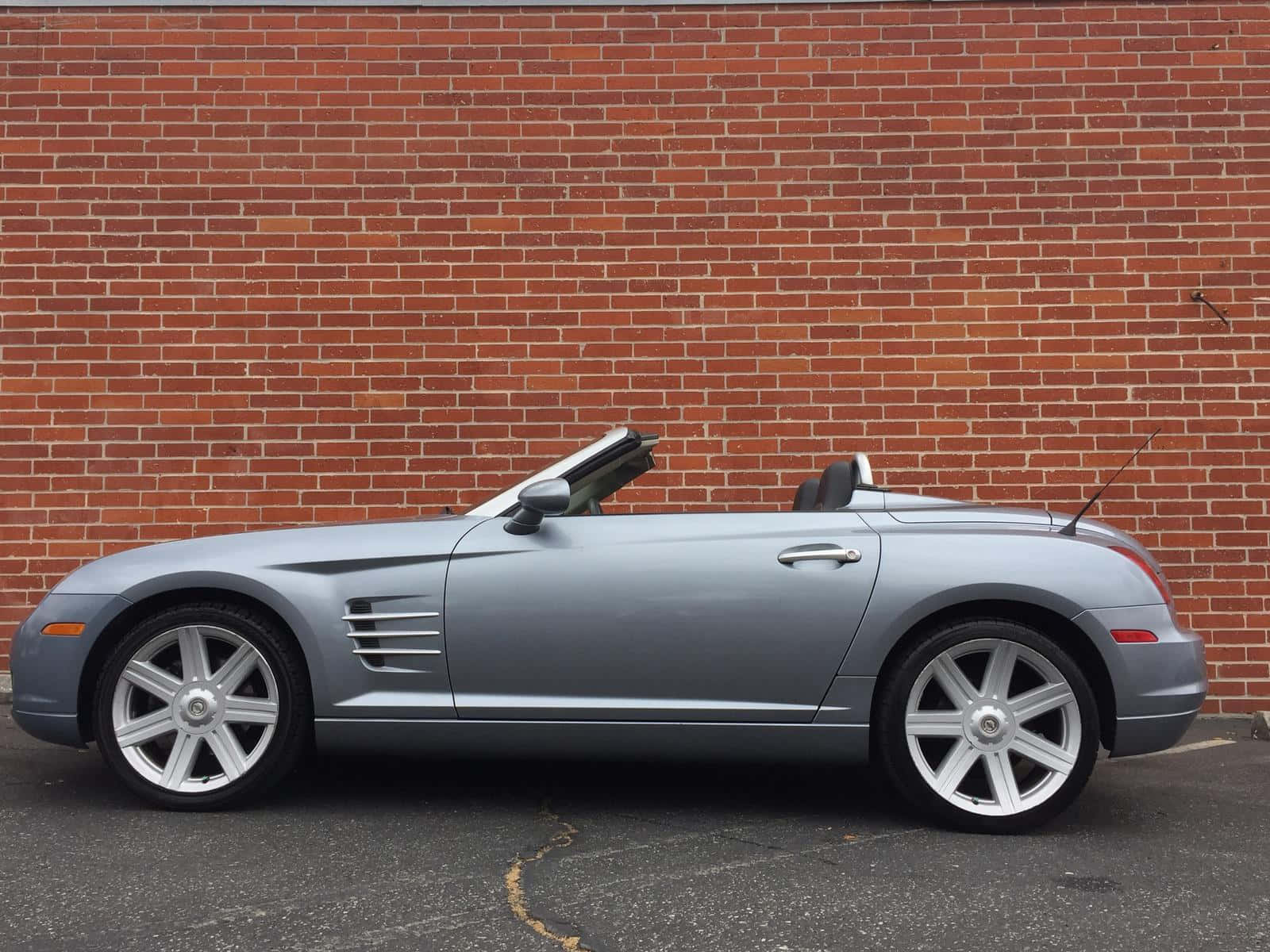 Chrysler Crossfire 桌布 壁紙 — 一架時尚的銀色開篷跑車停泊在紅磚牆前的瀝青路面上。