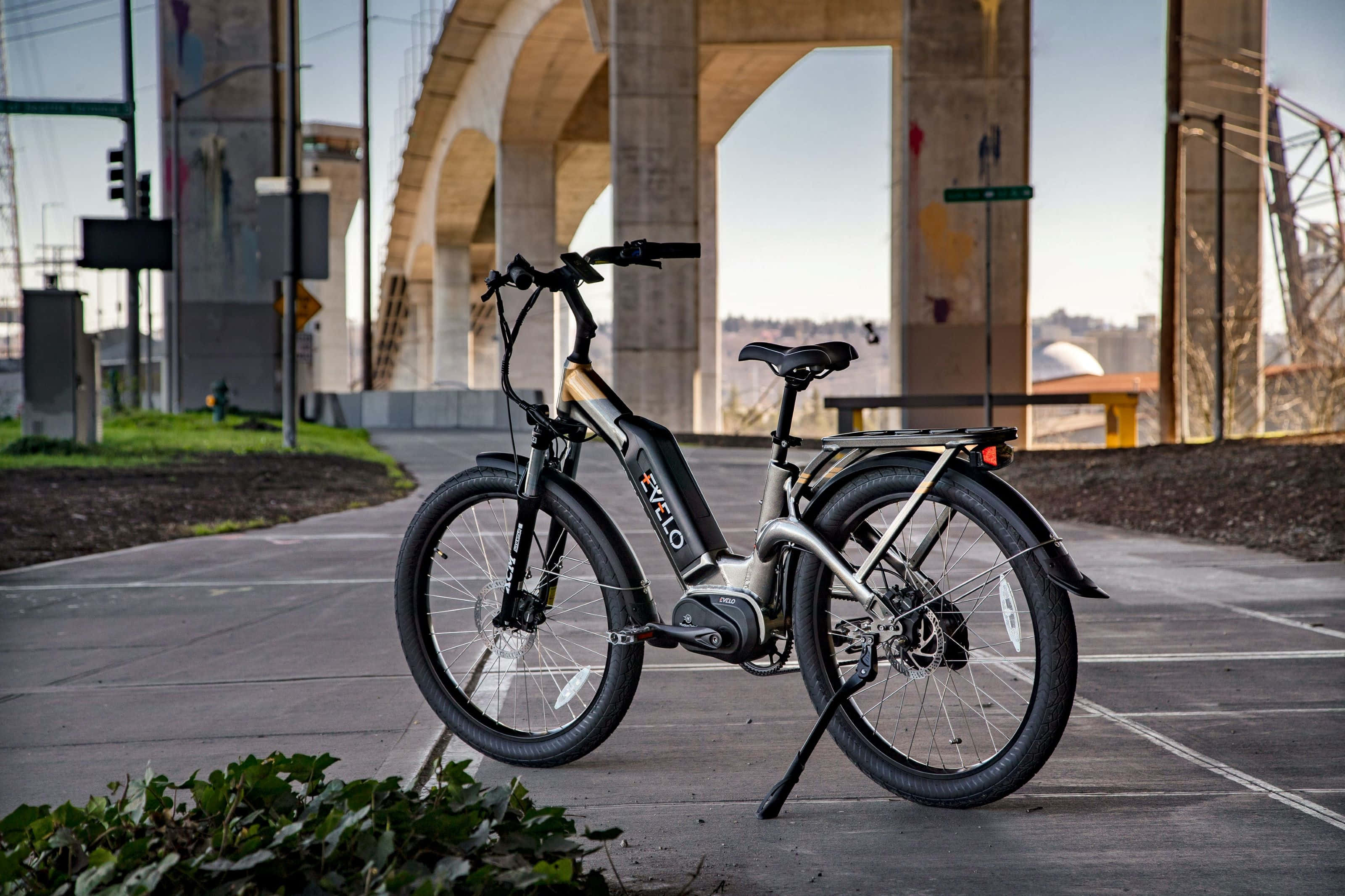 Electric Bike 桌布 壁紙 — 一架時尚的銀色電動單車停泊在混凝土橋下，橋柱上有塗鴉，附近有綠化。
