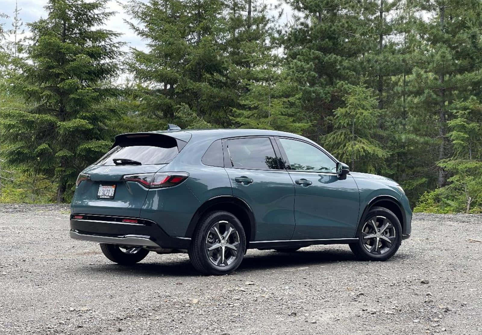 Honda Hr-v 桌布 壁紙 — 一架時尚的青綠色SUV停泊在碎石上，背景是高聳的常青樹，天空多雲。