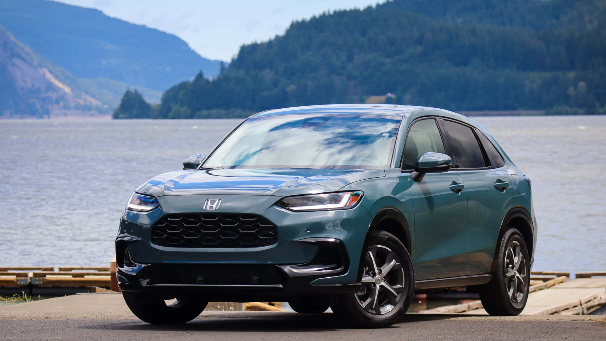Honda Hr-v 桌布 壁紙 — 一架時尚的青色本田SUV停泊在寧靜的湖邊，背景是森林覆蓋的山脈，天空晴朗。