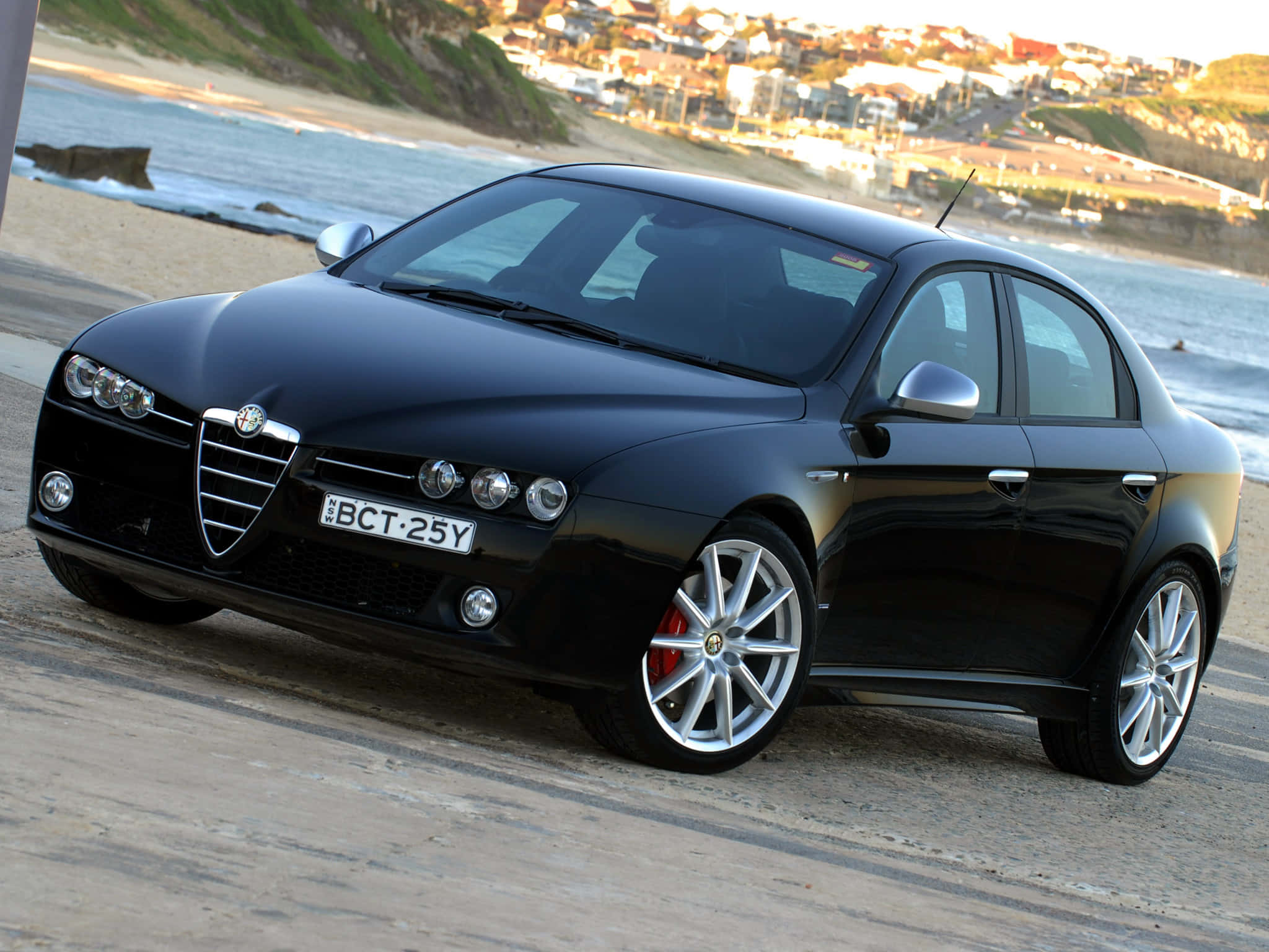 Alfa Romeo 159 桌布 壁紙 — 一架時尚的黑色 Alfa Romeo 房車停泊在沿海公路上，背景是海洋和懸崖小鎮。