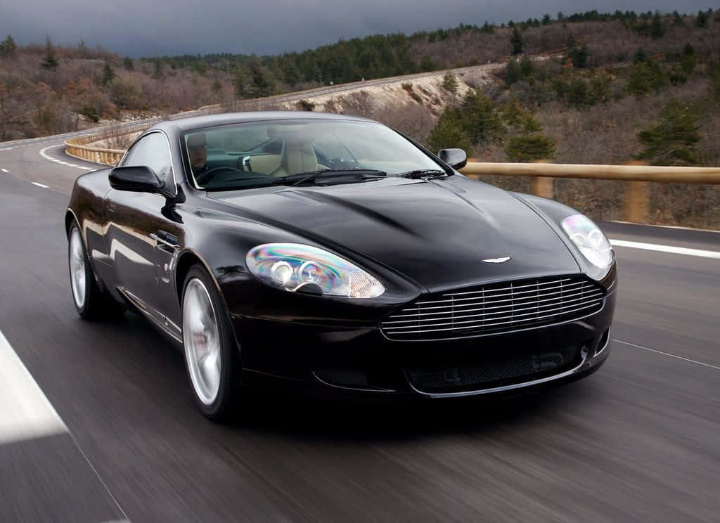 Aston Martin Db9 桌布 壁紙 — 一架時尚的黑色 Aston Martin 在陰天之下沿著蜿蜒的山路疾馳。
