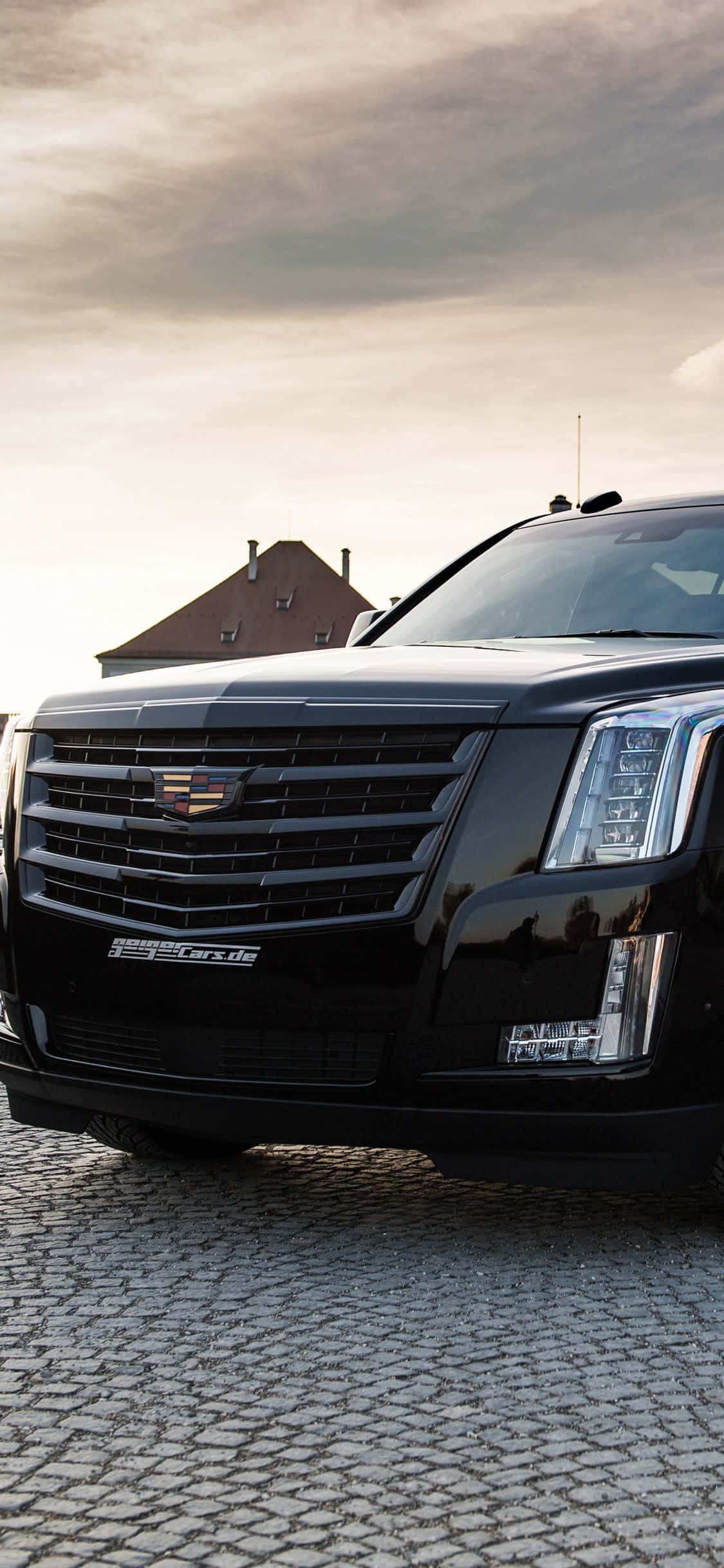 Cadillac Escalade 桌布 壁紙 — 一架時尚的黑色 Cadillac Escalade 停泊在鵝卵石街道上，天空多雲，背景有一間房屋。