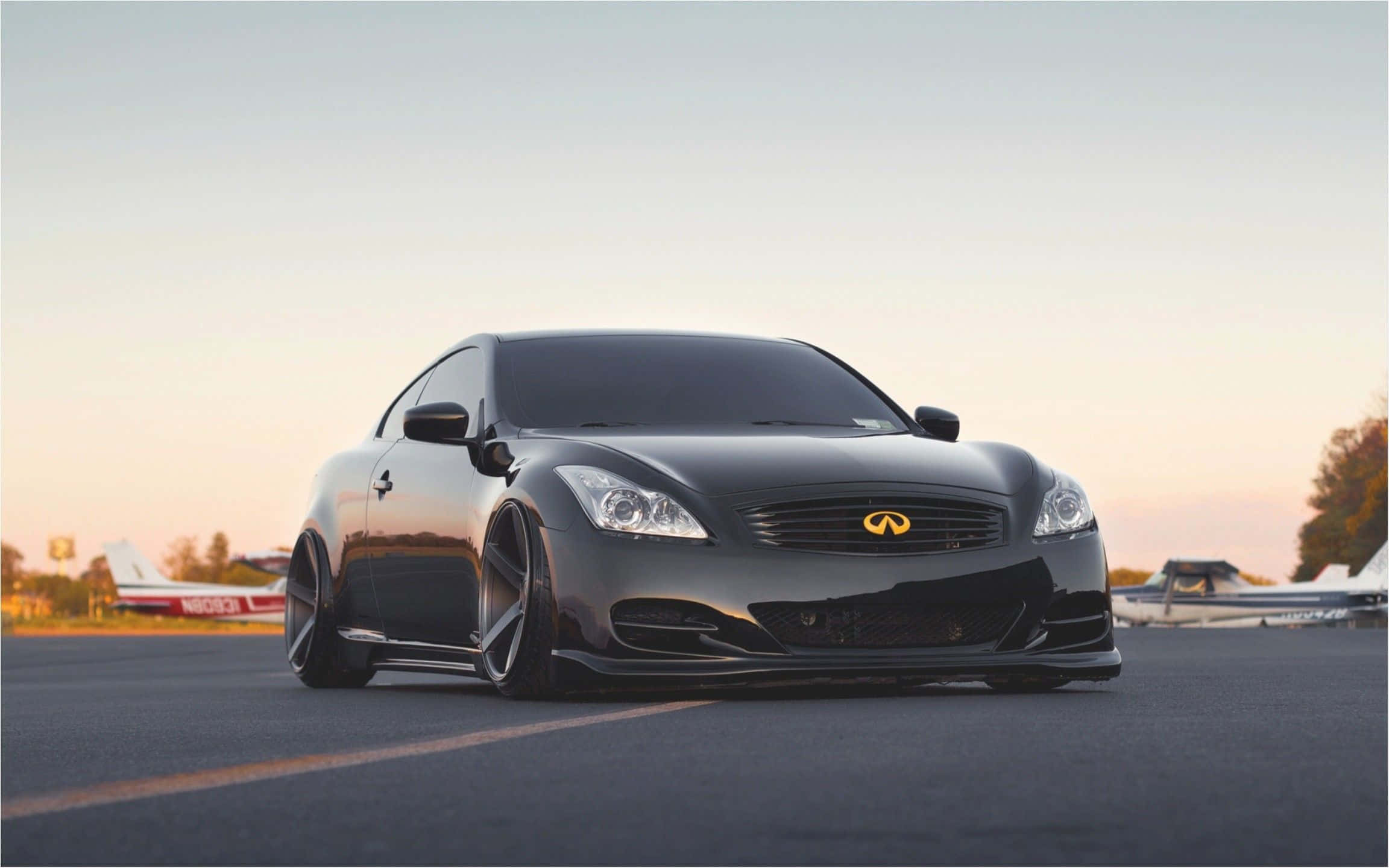 JDM 車 桌布 壁紙 — 一架時尚的黑色 Infiniti 雙門跑車配備定制輪圈，在黃金時段低趴於機場停機坪上，背景有小飛機。