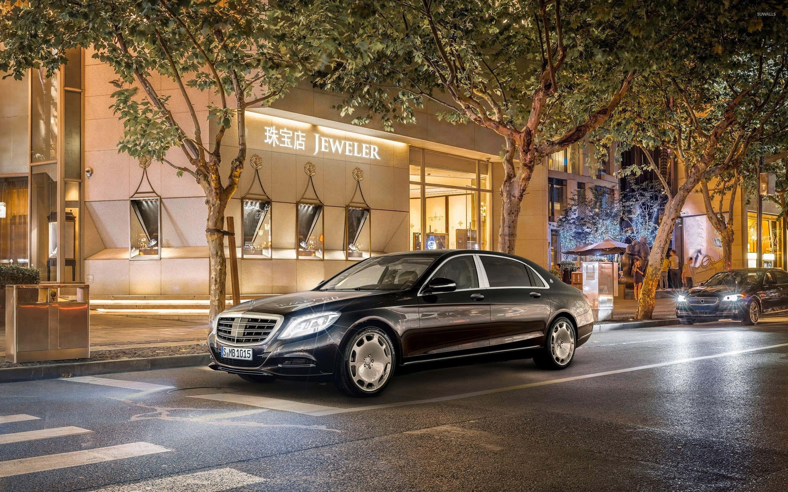 Maybach 桌布 壁紙 — 一架時尚的黑色 Mercedes-Maybach S-Class 在夜晚沿著濕漉漉的城市街道滑行，經過一間閃閃發光的珠寶店。