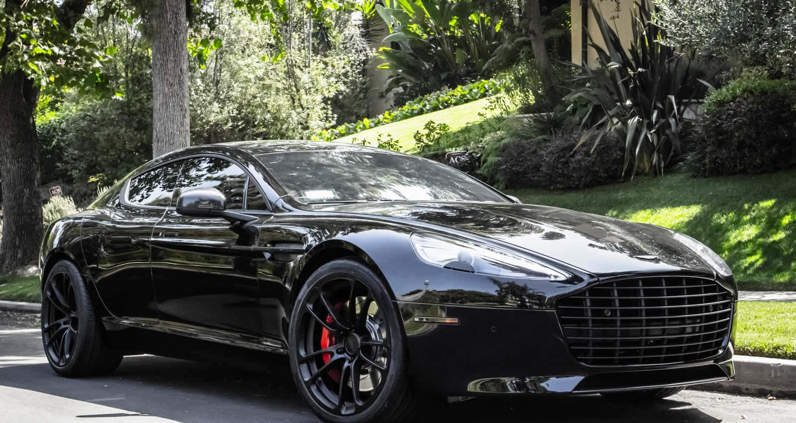 Aston Martin Rapide S 桌布 壁紙 — 一架時尚的黑色Aston Martin跑車在明亮的陽光下閃閃發光，停泊在綠樹成蔭的住宅街道上。