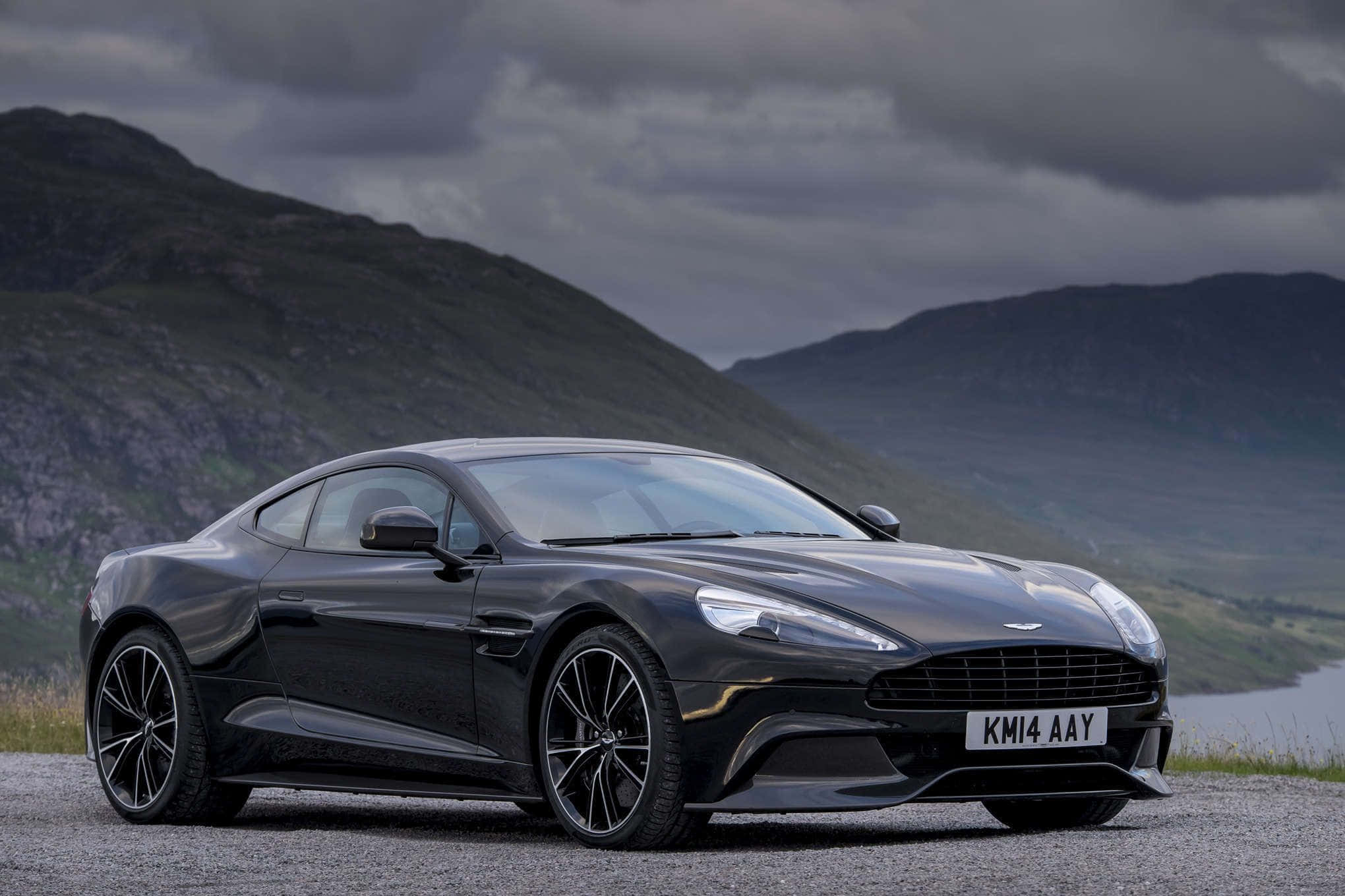 Aston Martin Vanquish 桌布 壁紙 — 一架時尚的黑色Aston Martin跑車停泊在碎石路上，背景係戲劇性嘅山區地形同多雲嘅天空。