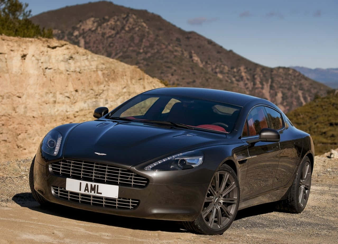 Aston Martin Rapide S 桌布 壁紙 — 一架時尚的黑色Aston Martin跑車停泊在塵土飛揚的山路上，背景是崎嶇的山丘和清澈的藍天。