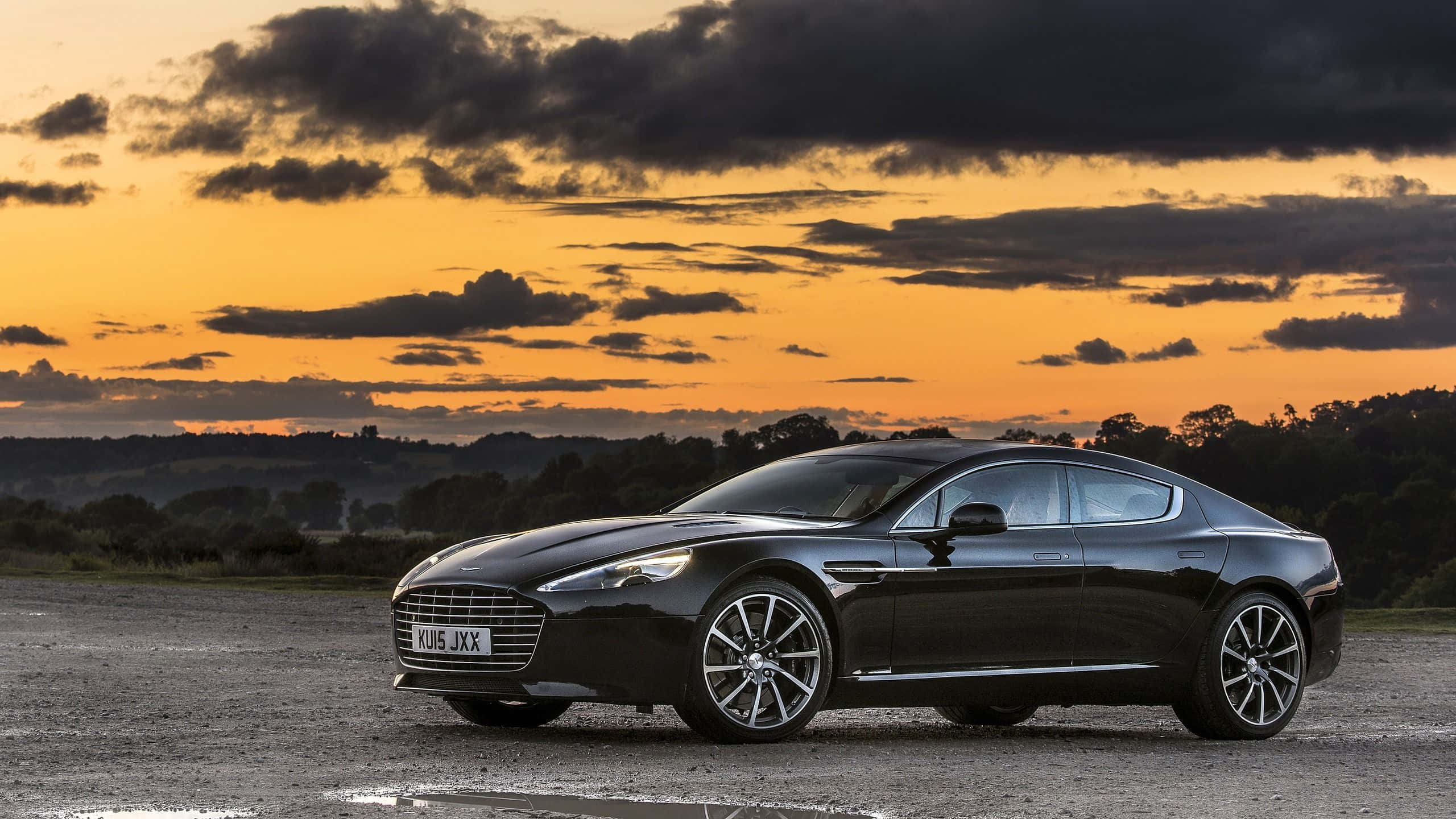 Aston Martin Rapide S 桌布 壁紙 — 一架時尚的黑色Aston Martin Rapide停泊在碎石地上，背景是帶有橙色雲彩的戲劇性日落天空。