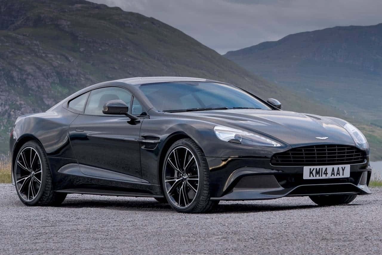 Aston Martin Vanquish 桌布 壁紙 — 一架時尚的黑色Aston Martin跑車停泊在碎石上，背景是陰天多雲的天空下霧氣瀰漫的山脈。