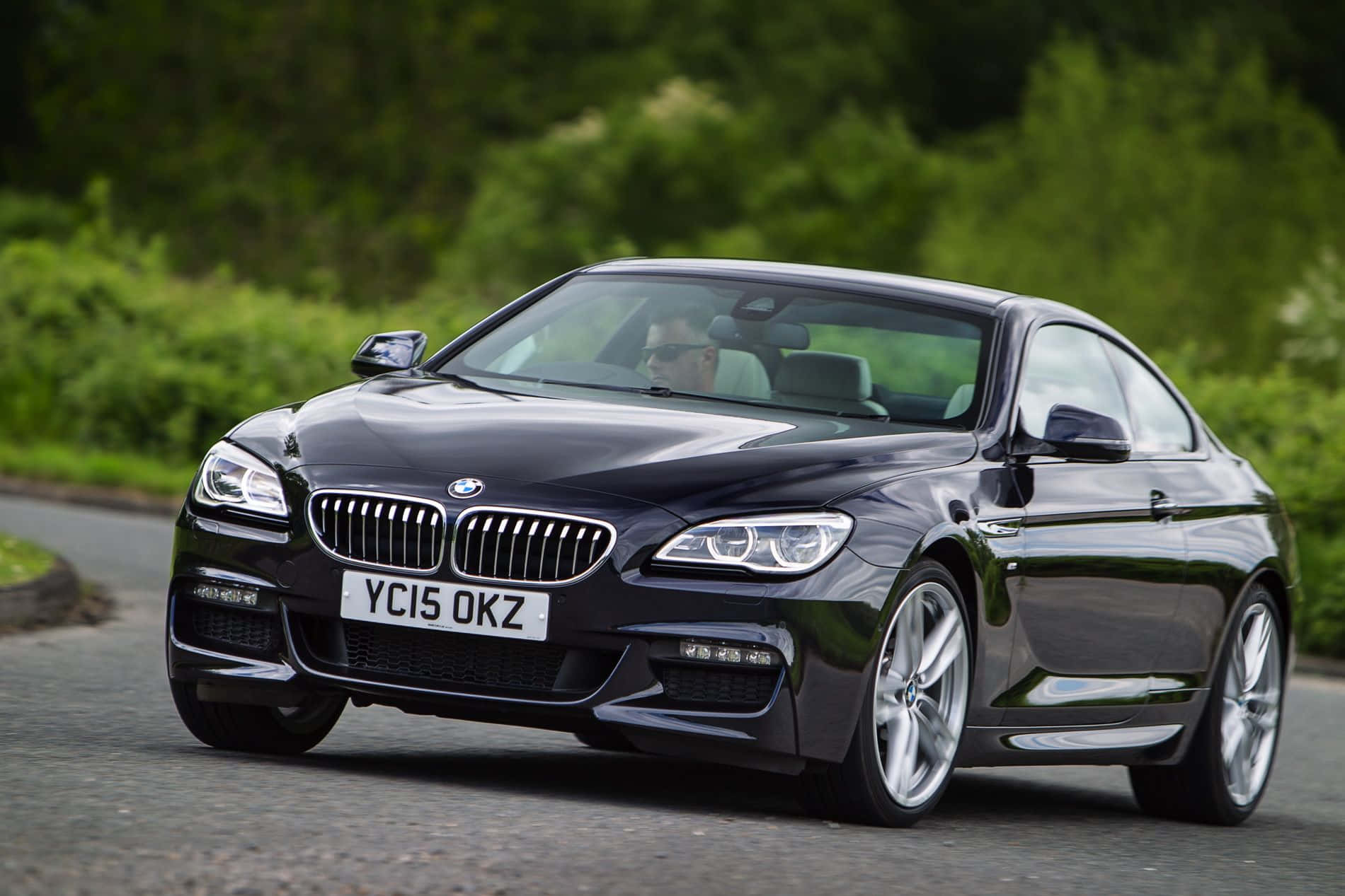 Bmw 6 Series 桌布 壁紙 — 一架時尚的黑色BMW 6系轎跑車沿著蜿蜒的道路行駛，周圍環繞著茂密的綠色樹葉。