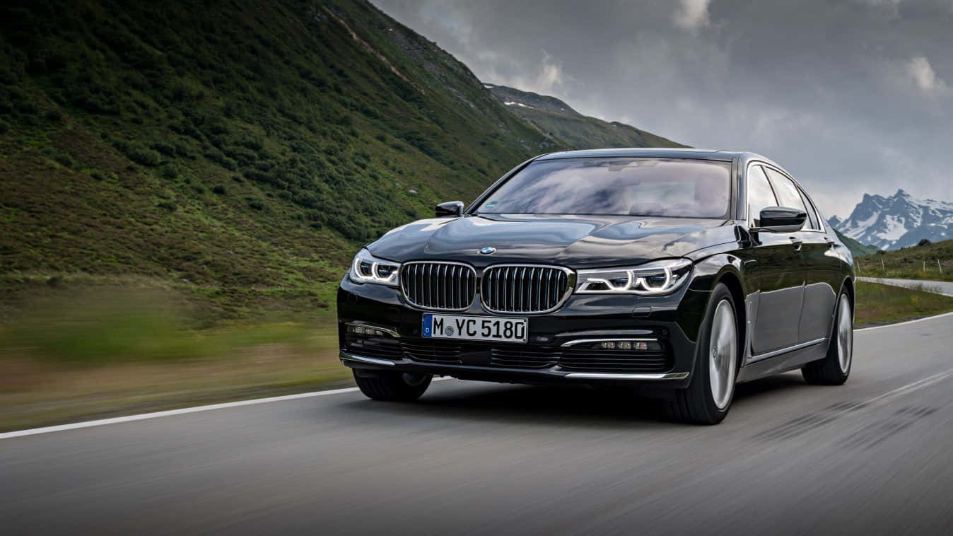 Bmw 7 Series 桌布 壁紙 — 一架時尚的黑色BMW 7系房車在蜿蜒的山路上疾馳，兩旁是翠綠的山坡和積雪的山峰，天空多雲。
