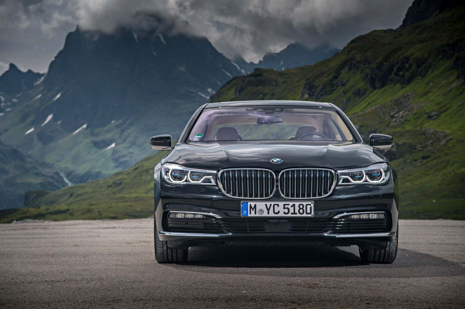 Bmw 7 Series 桌布 壁紙 — 一架時尚的黑色BMW 7系轎車在山上道路向前行駛，背後有戲劇性的綠色山峰和暴風雲。