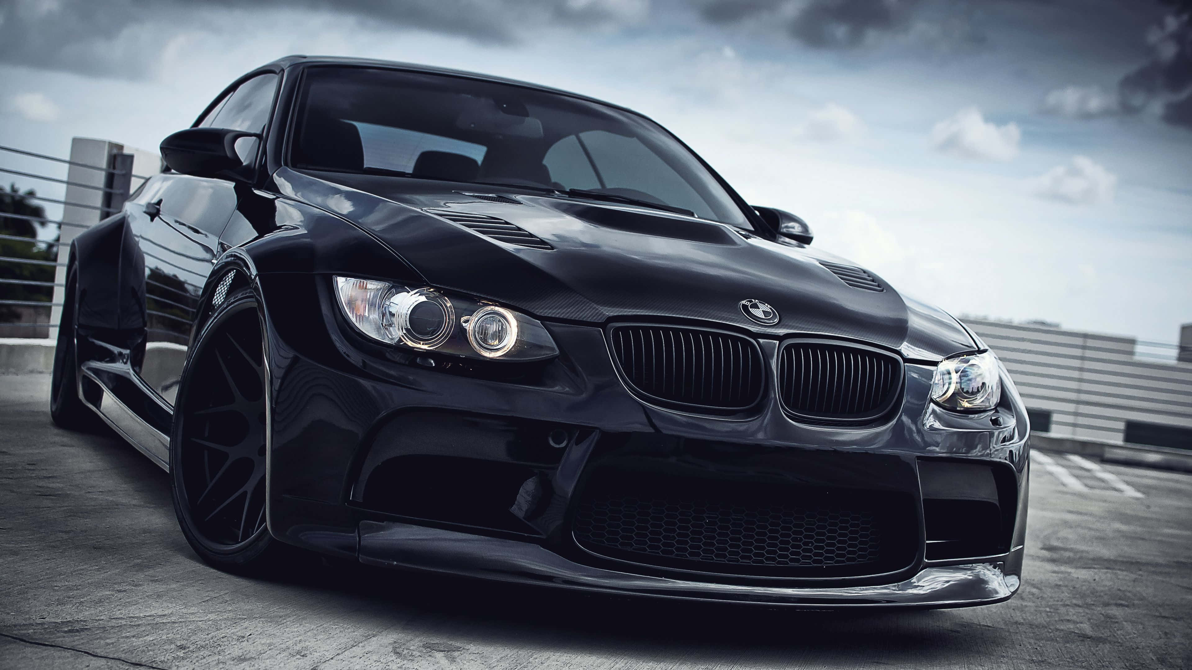 4K BMW 壁紙 桌布 壁紙 — 一架時尚的黑色BMW M3跑車，配備自訂寬體套件和大輪圈，停泊在混凝土屋頂上，天空多雲。