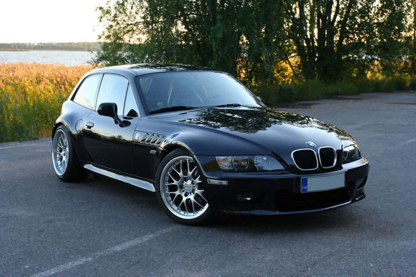 Bmw Z3 桌布 壁紙 — 一架時尚的黑色BMW Z3跑車在黃金時段停泊在近水邊的高草和樹木旁的瀝青路上。