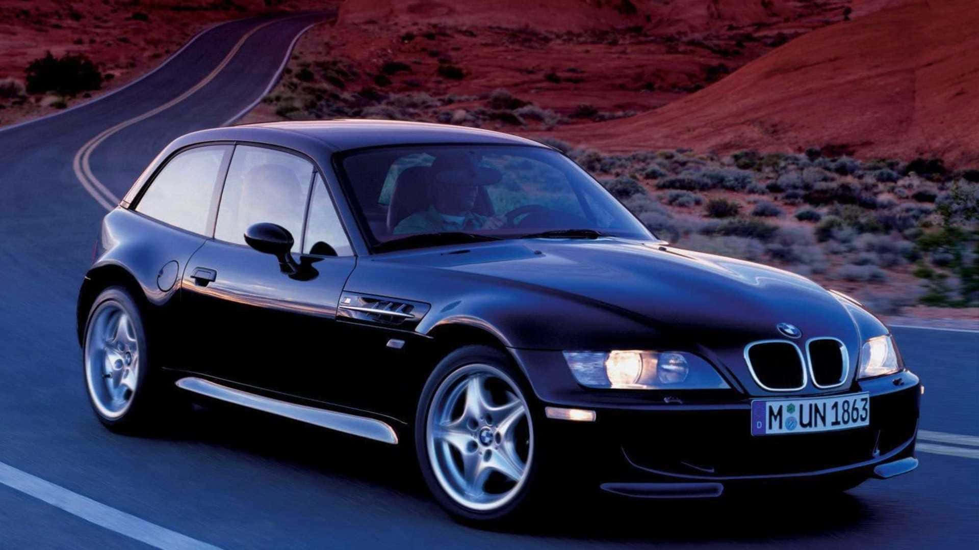Bmw Z3 桌布 壁紙 — 一架時尚的黑色BMW Z3跑車在日落時分沿著蜿蜒的沙漠公路行駛，背景是紅色岩層。
