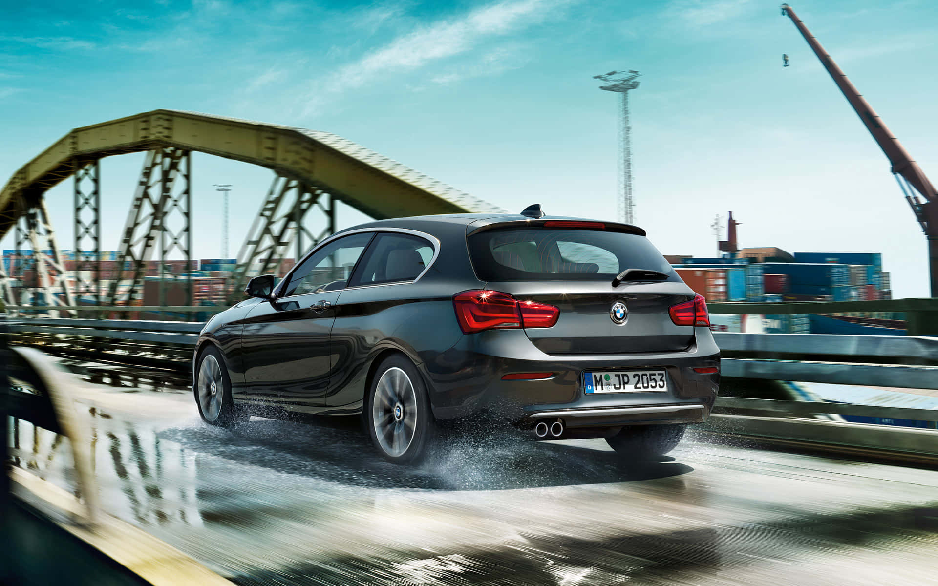 Bmw 1 Series 桌布 壁紙 — 一架時尚的黑色BMW掀背車在濕滑的橋上飛馳，背景是貨運集裝箱和起重機，天空湛藍清澈。