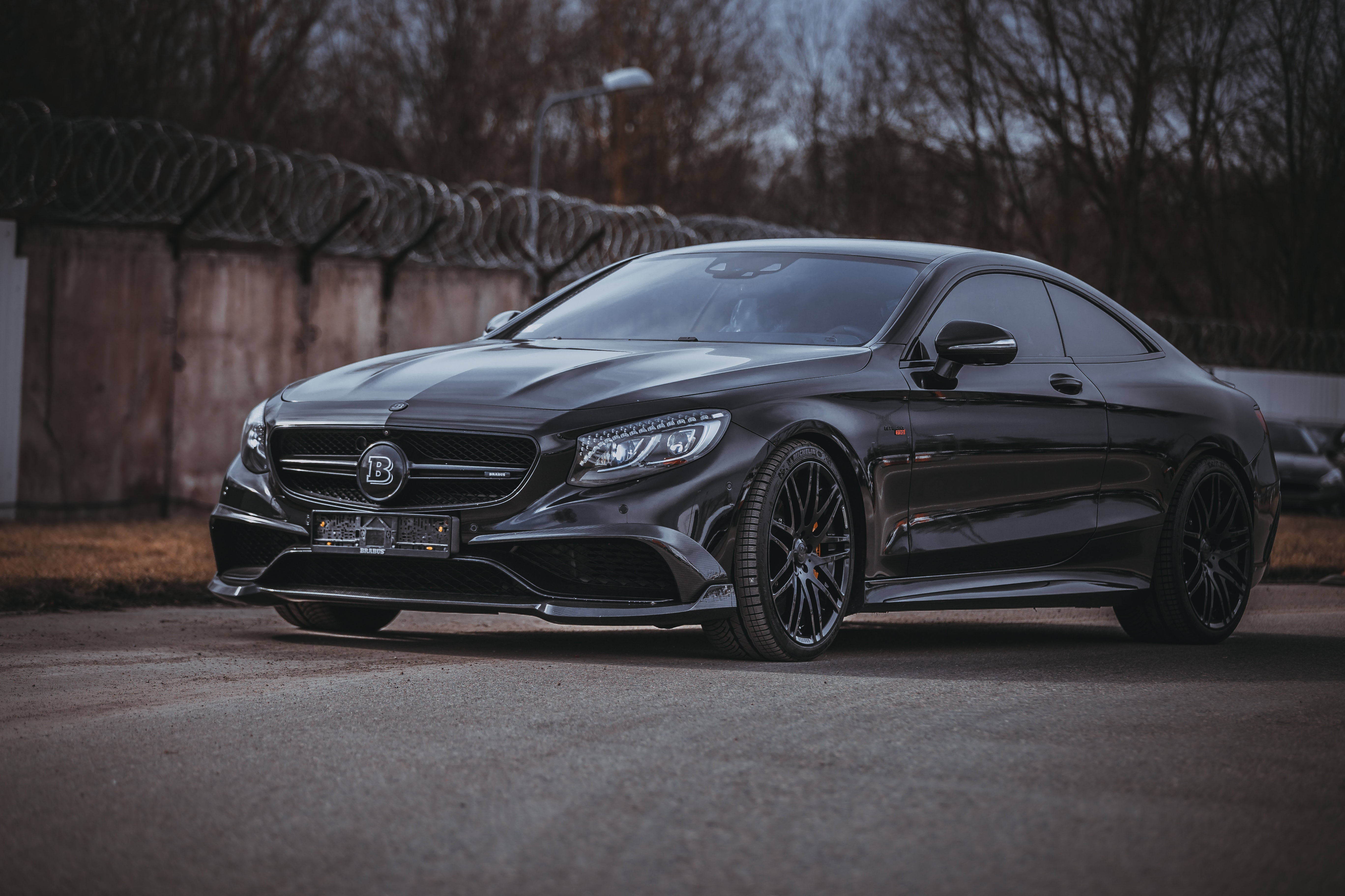 HD 桌布 壁紙 — 一架時尚的黑色Brabus Mercedes-AMG S 65 Coupe停泊在瀝青路上，靠近帶刺鐵絲網圍欄，天空多雲。
