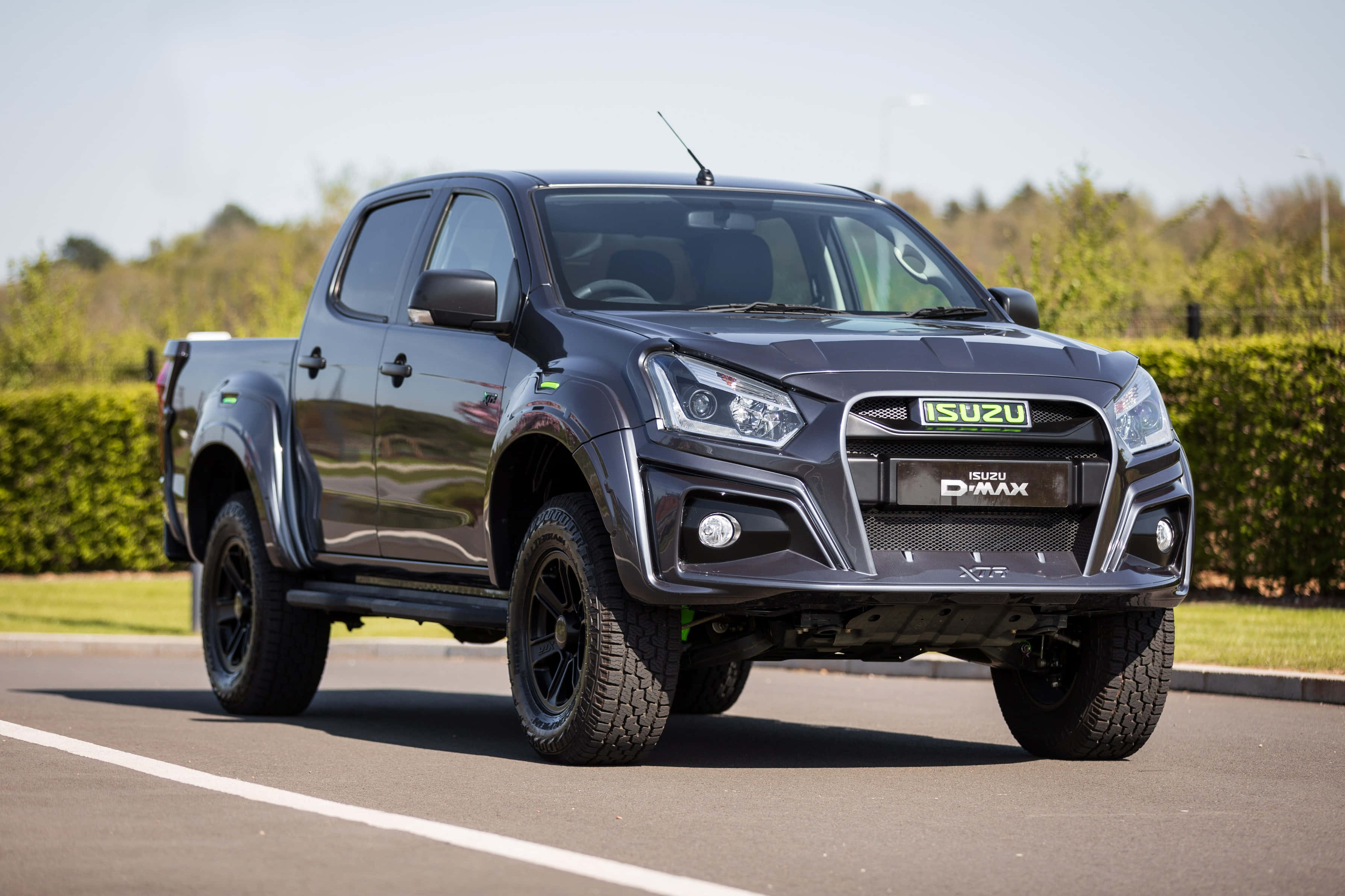 Isuzu 桌布 壁紙 — 一架時尚的黑色Isuzu D-MAX皮卡車停在瀝青路上，背景有綠色籬笆和樹木，天空晴朗。