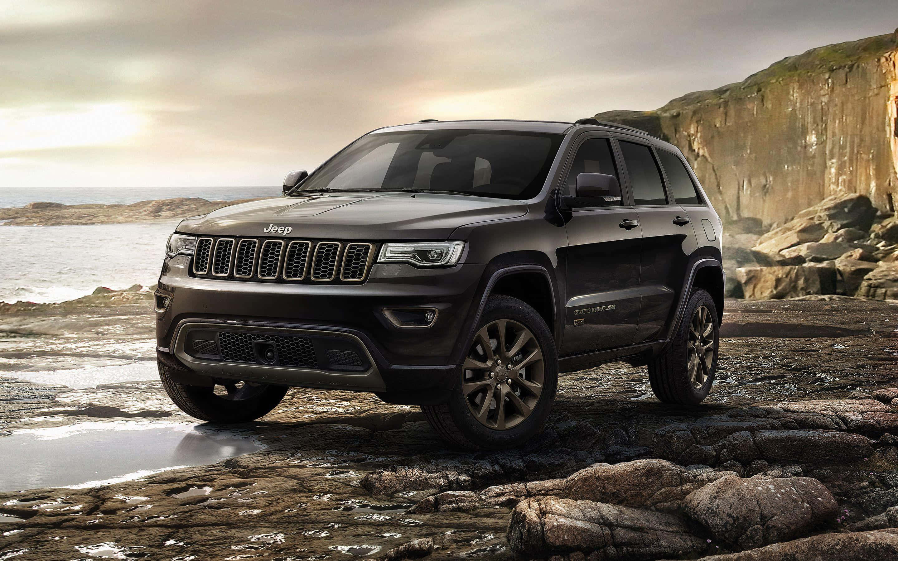 Jeep Wagoneer 桌布 壁紙 — 一架時尚的黑色Jeep Grand Cherokee停在濕漉漉的海岸岩石上，背景是戲劇性的懸崖和海洋，天空多雲。