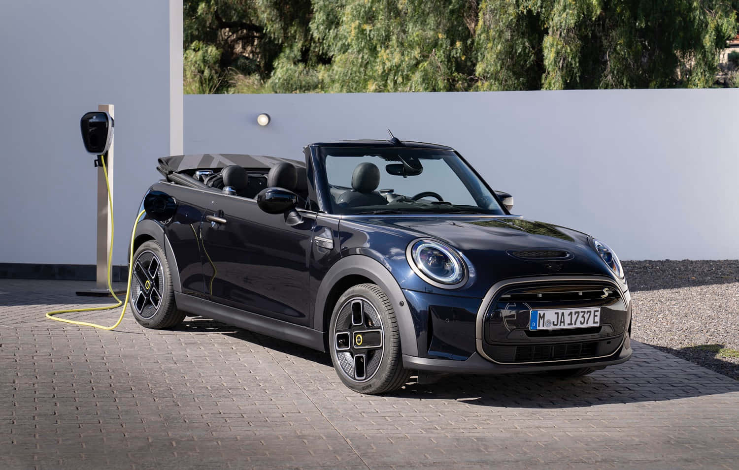 Mini Cooper S Convertible 桌布 壁紙 — 一架時尚的黑色MINI Cooper開篷車在陽光明媚的日子裡，在白色牆壁後有綠樹的充電站充電。