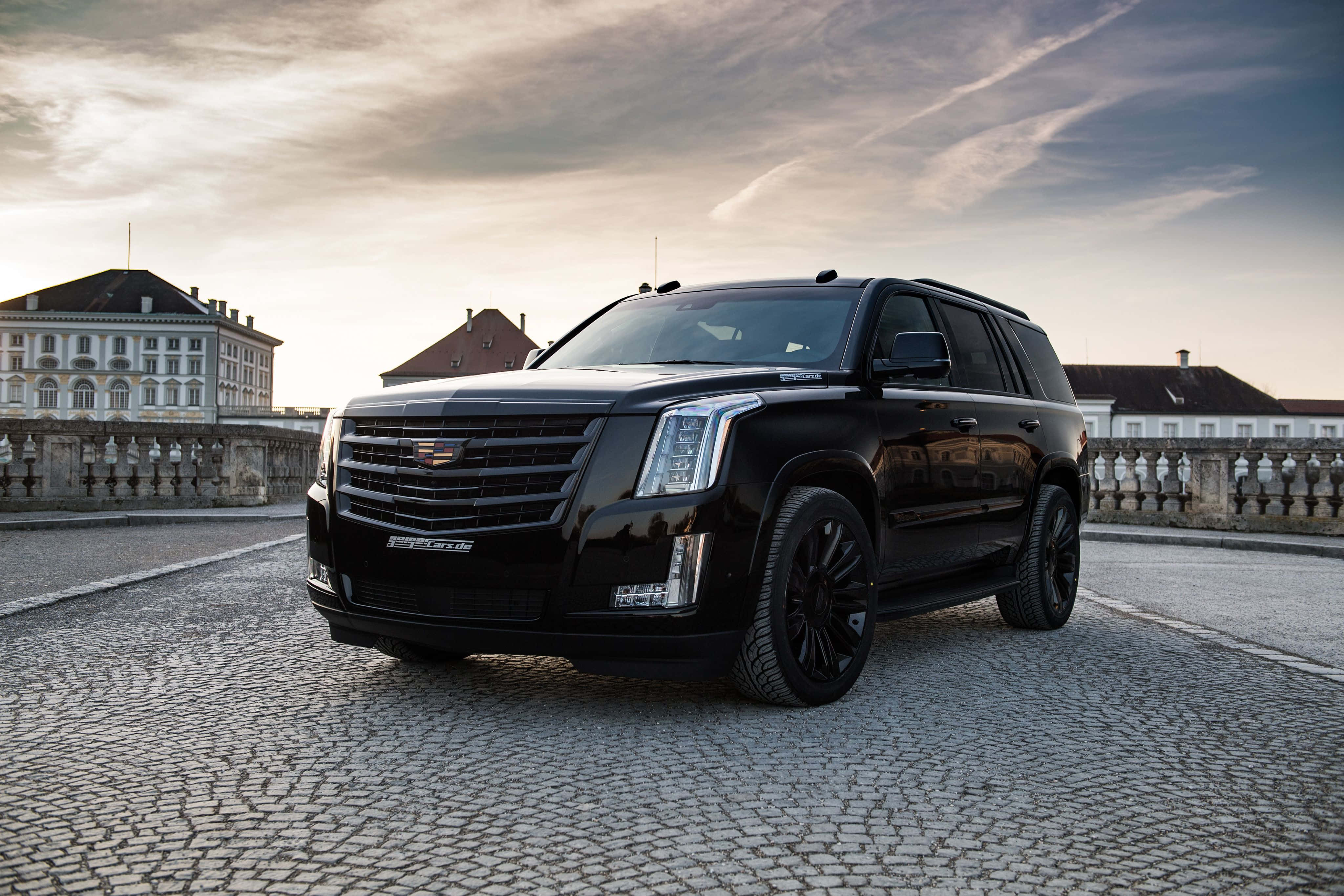 Cadillac Escalade 桌布 壁紙 — 一架時尚的黑色凱迪拉克 Escalade 停泊在鵝卵石露台上，背景是歷史建築，天空呈現柔和的金色日落。