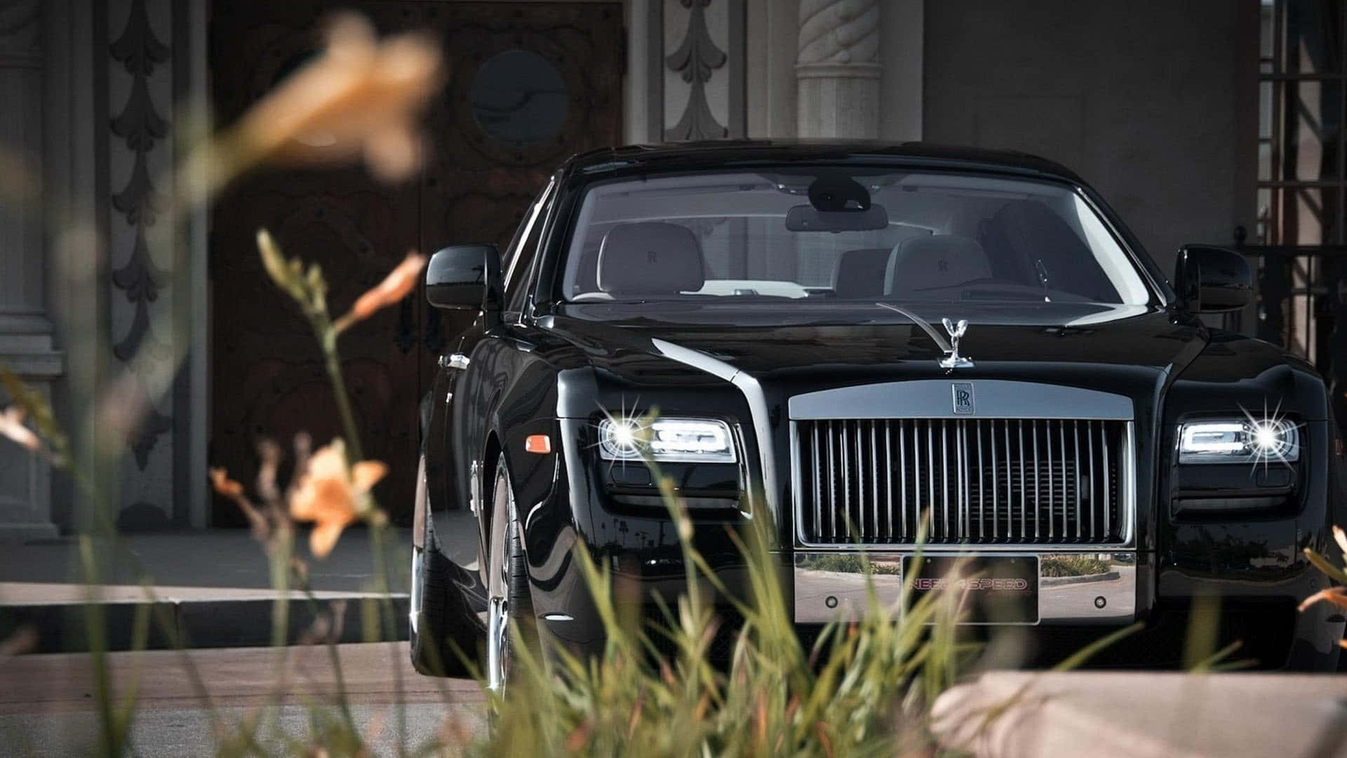 Rolls Royce 桌布 壁紙 — 一架時尚的黑色勞斯萊斯幻影在日光下閃耀，以華麗的建築和前景模糊的橙色花朵作為框架。