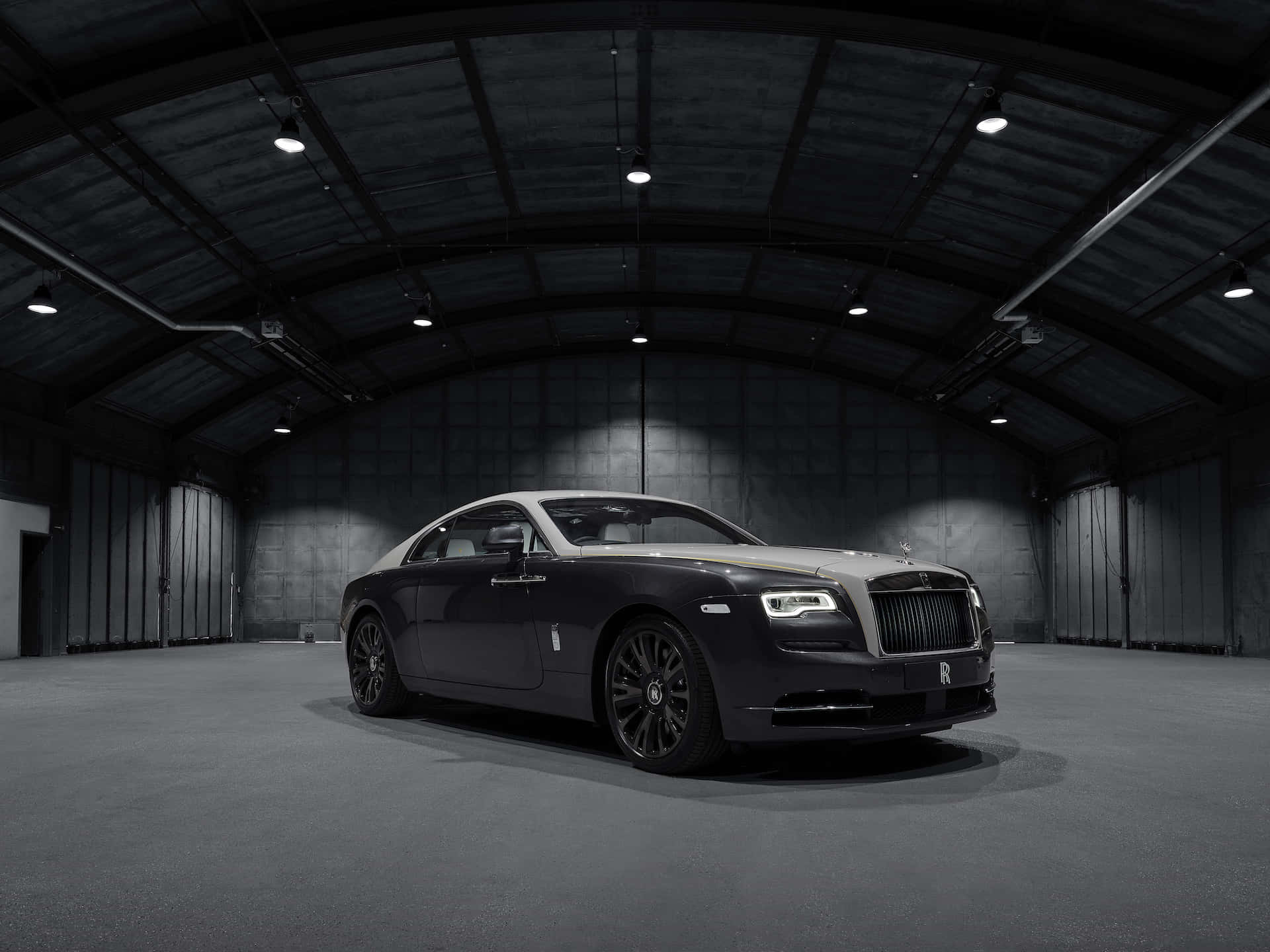 Rolls Royce 桌布 壁紙 — 一架時尚的黑色勞斯萊斯魅影在寬敞的工業機庫內，高拱形天花板的聚光燈下閃閃發光。