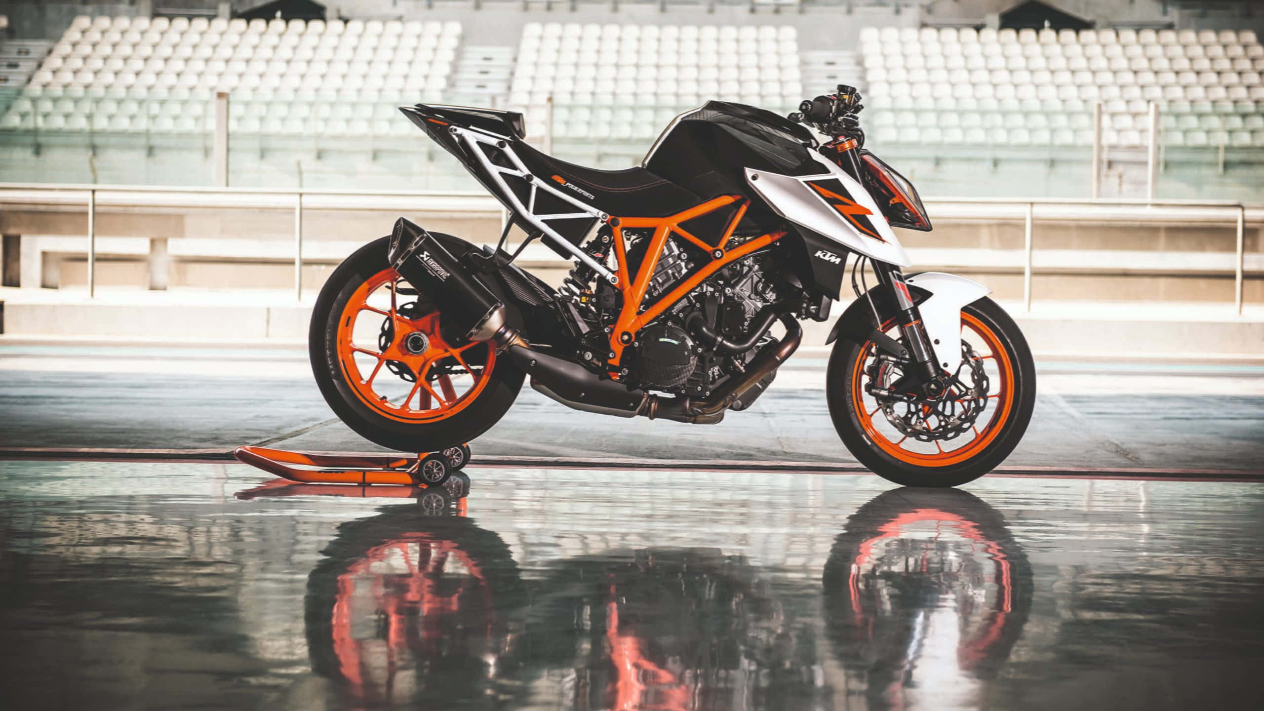 Ktm Bike 桌布 壁紙 — 一架時尚的黑色和橙色KTM電單車停在一個空蕩蕩的體育館內，地面濕滑反光，周圍是白色座位。