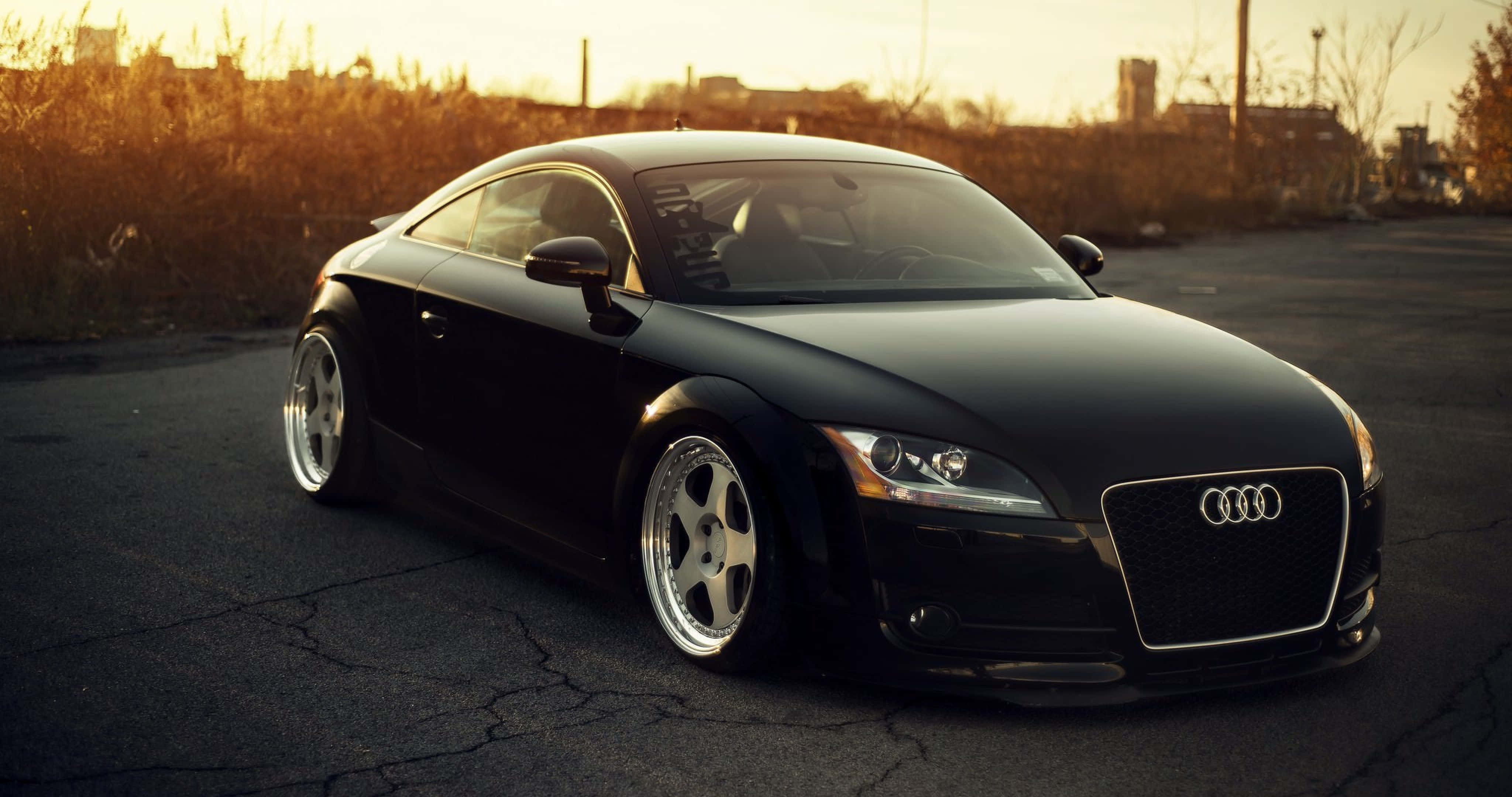 Audi Tt 桌布 壁紙 — 一架時尚的黑色奧迪TT雙門跑車停泊在龜裂的瀝青路上，沐浴在溫暖的金色夕陽光中。