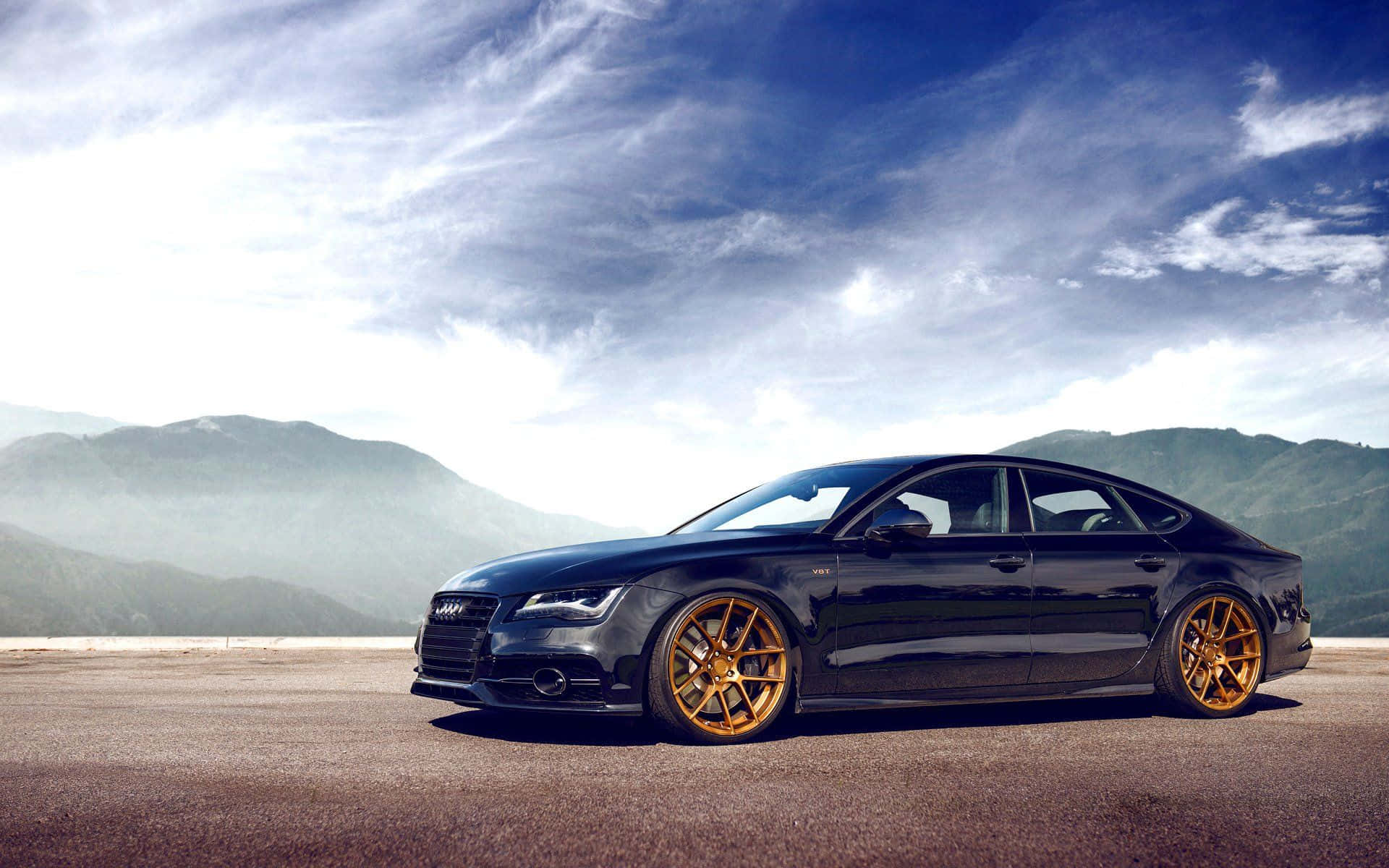 Audi S7 桌布 壁紙 — 一架時尚的黑色奧迪轎車配金色輪圈，停泊在山頂觀景點，背景是戲劇性的多雲天空。