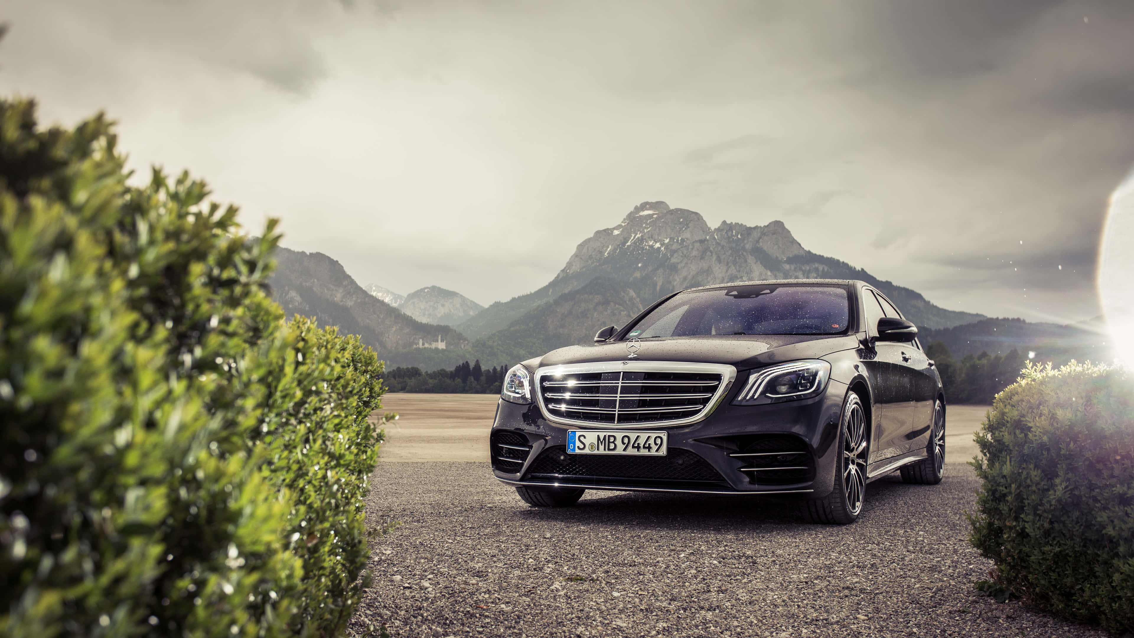 4K Mercedes 桌布 壁紙 — 一架時尚的黑色平治S-Class房車停泊在碎石路上，兩旁是綠色籬笆，背景是雲天之下積雪的山峰。