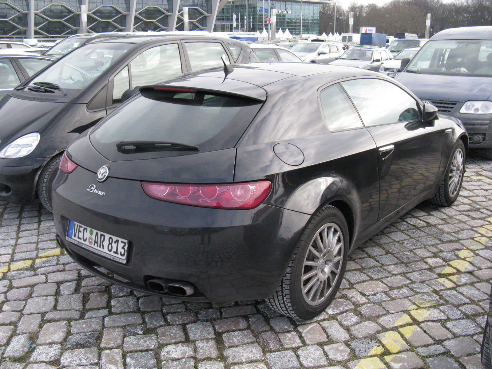 Alfa Romeo Brera 桌布 壁紙 — 一架時尚的黑色愛快羅密歐 Brera 雙門跑車停泊在鵝卵石停車場，背景是現代建築。