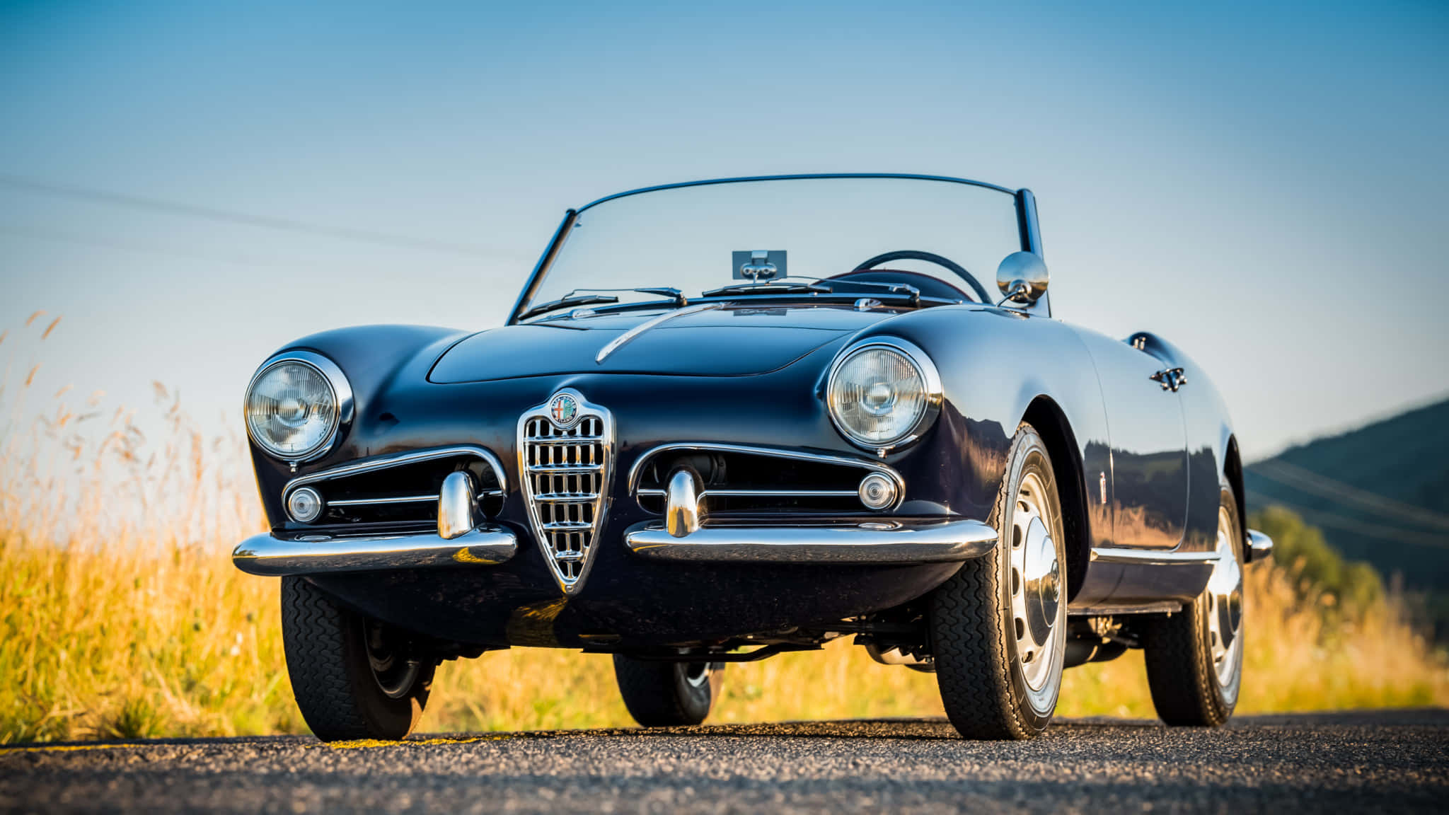 Alfa Romeo Spider 桌布 壁紙 — 一架時尚的黑色愛快羅密歐Spider開篷車停泊在陽光明媚的鄉村道路上，周圍是金色的草地和遠處的山丘，天空清澈湛藍。