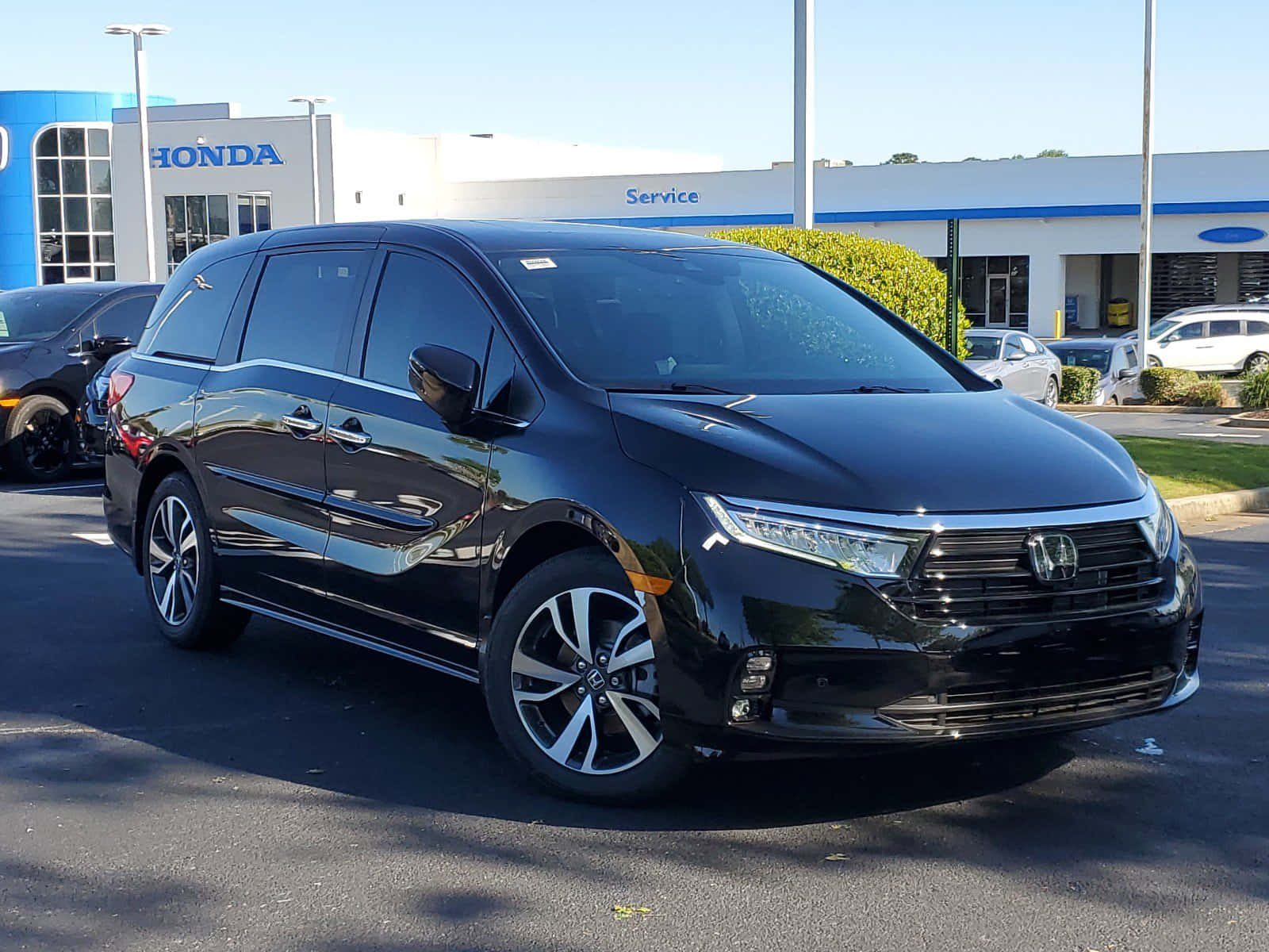 Honda Odyssey 桌布 壁紙 — 一架時尚的黑色本田小型客車在明亮的陽光下，於經銷商場地中閃閃發光，背景有其他汽車和服務大樓。