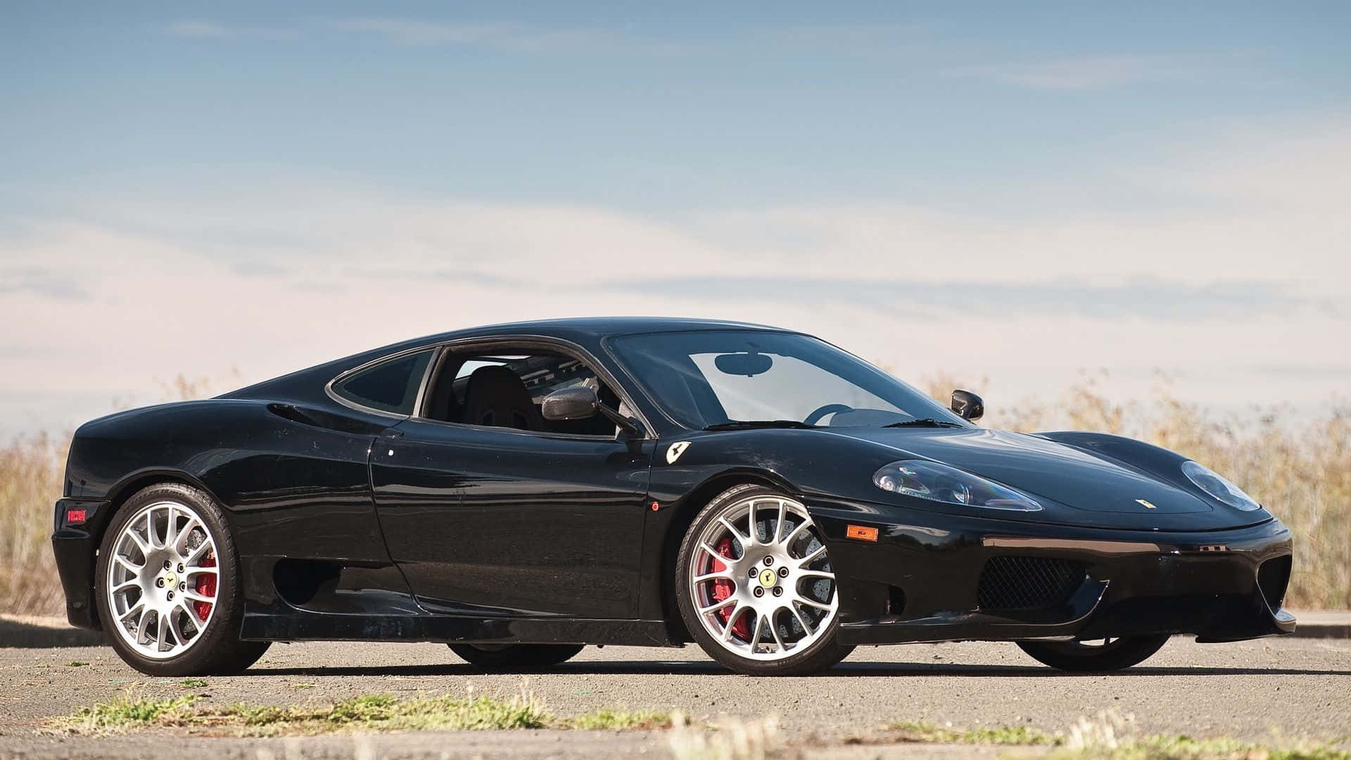 Ferrari 360 Modena 桌布 壁紙 — 一架時尚的黑色法拉利 360 跑車停泊在鋪砌的道路上，背景有乾草和晴朗的藍天。