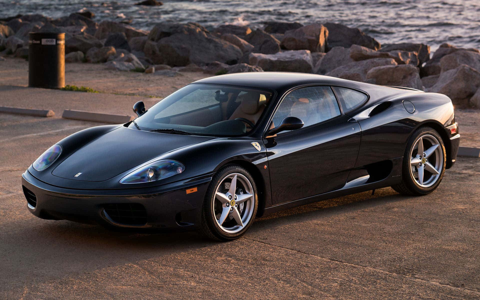 Ferrari 360 Modena 桌布 壁紙 — 一架時尚的黑色法拉利360跑車停泊在黃金時段岩石海岸線旁的鋪砌停車場上。