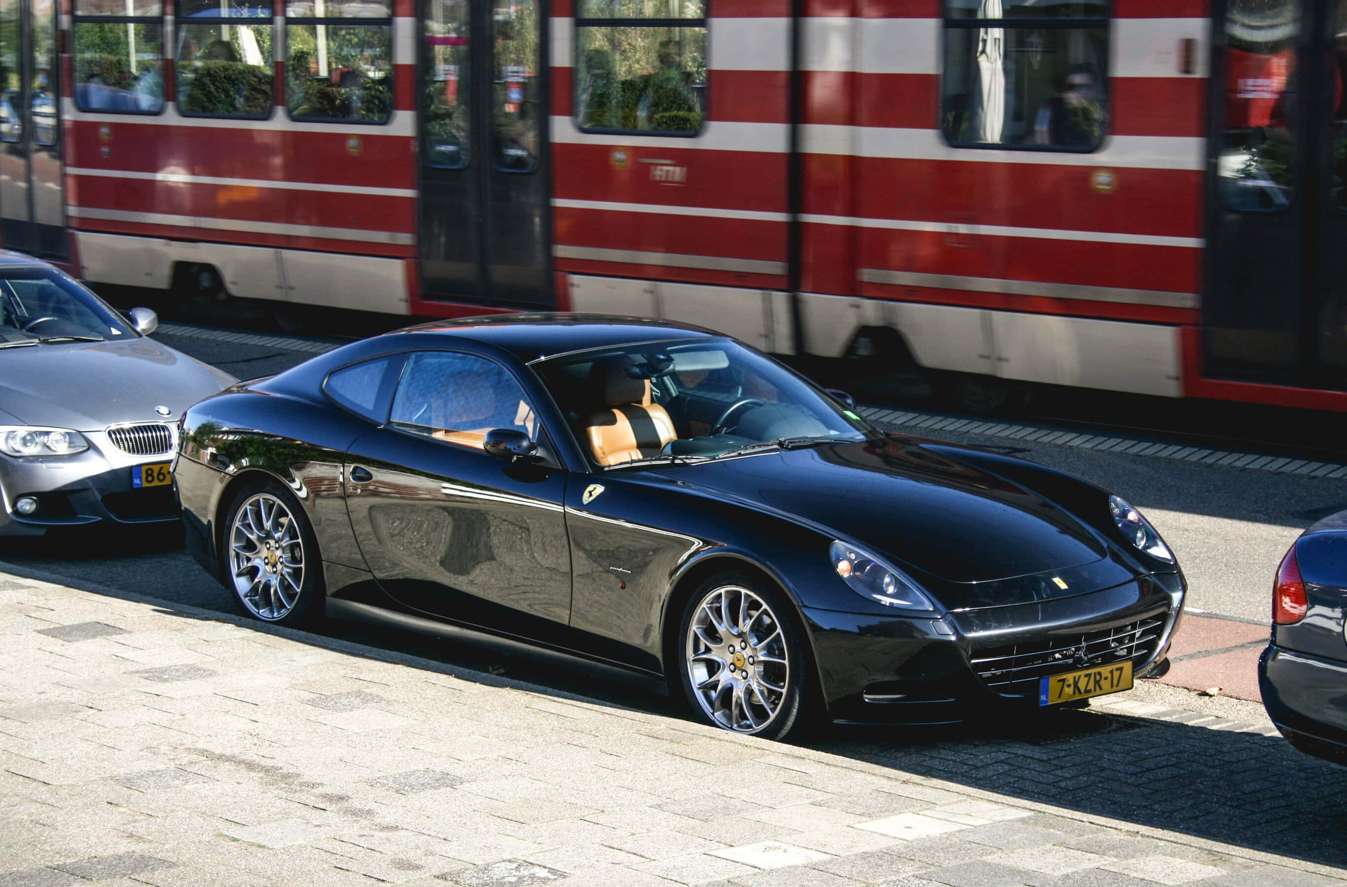 Ferrari 612 Scaglietti 桌布 壁紙 — 一架時尚的黑色法拉利California停泊在陽光普照的鵝卵石街道上，旁邊是一列紅白相間的火車。